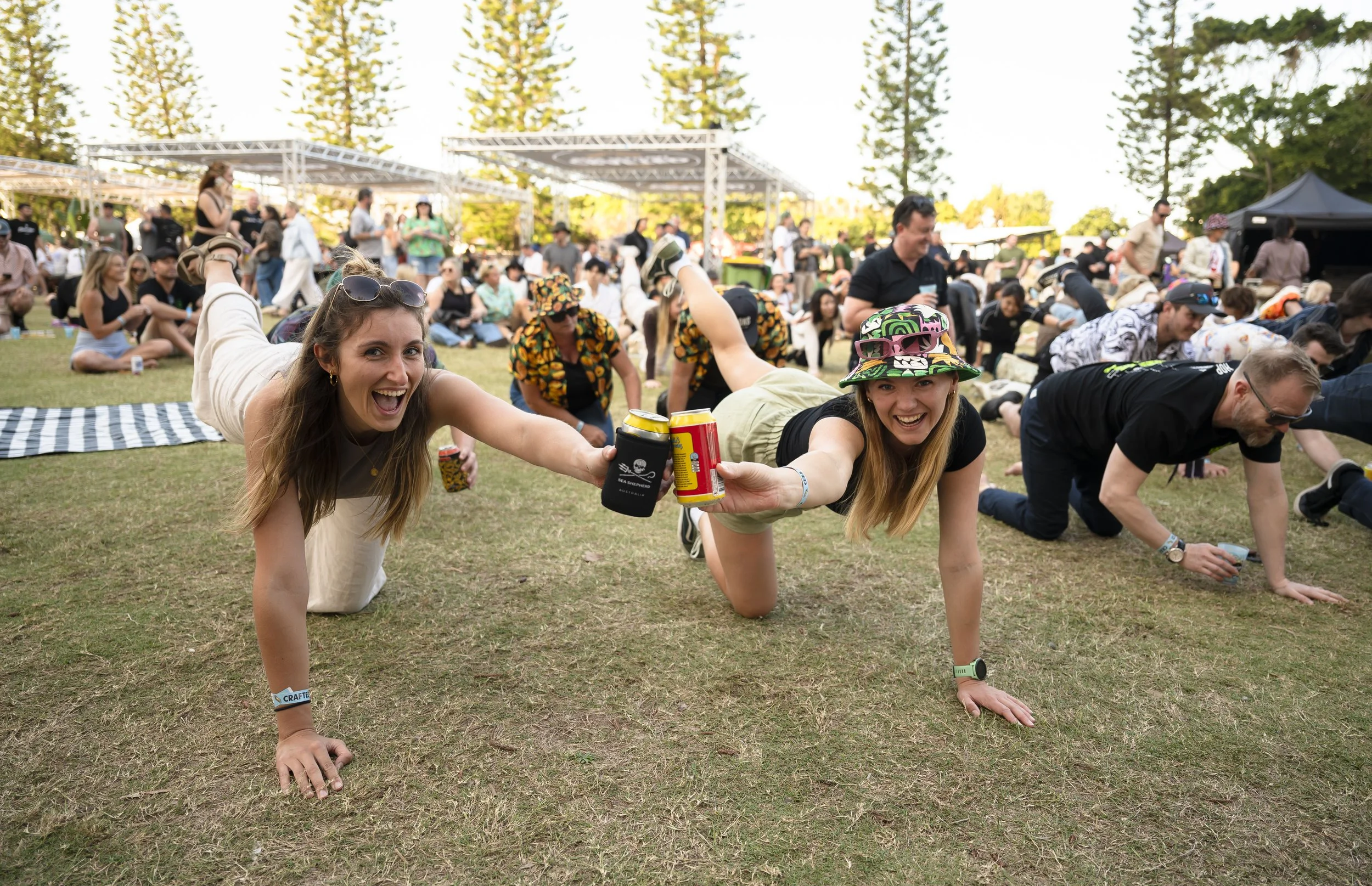 Two women doing a plank pose on the grass at an outdoor festival, holding drinks, with a large crowd in the background.