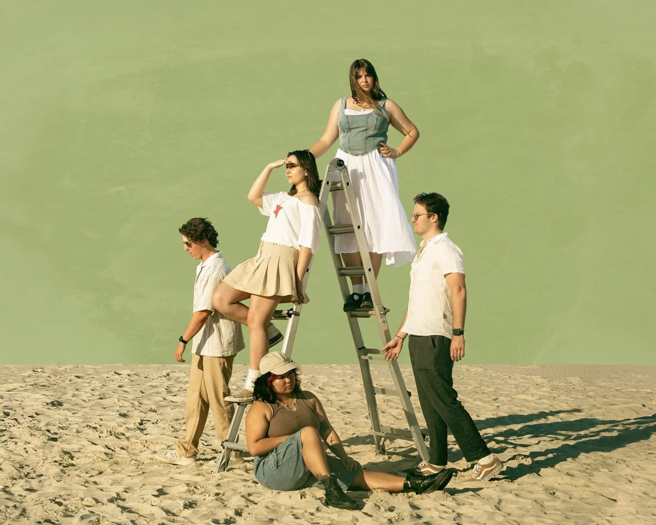 Group of six young people on the beach, some standing, some seated or on a ladder, against a green sky background, posing for a photo.