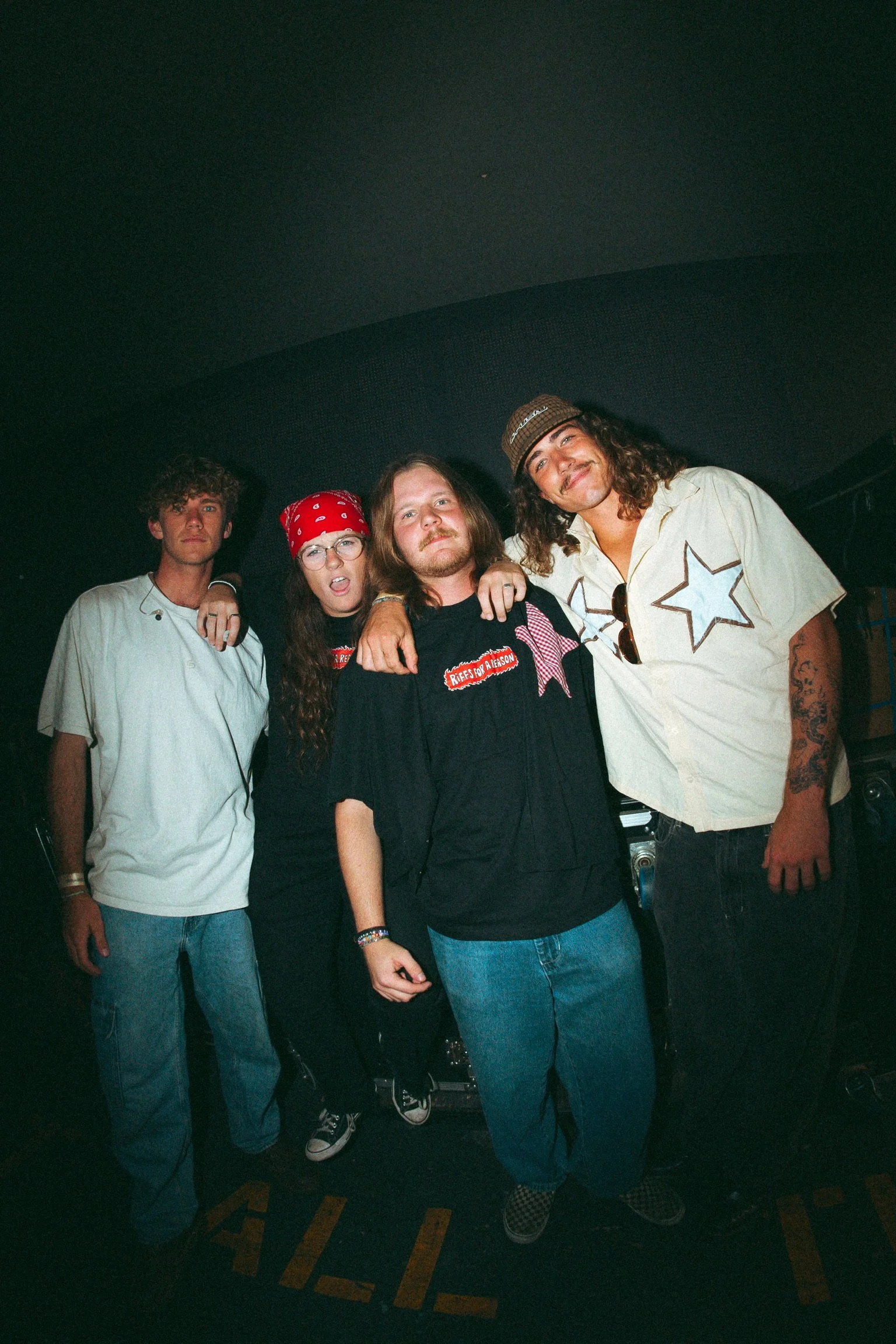 Group of four young people standing together in a dark setting, smiling and posing for the photo. They are dressed casually and appear to be at a social event or gathering.
