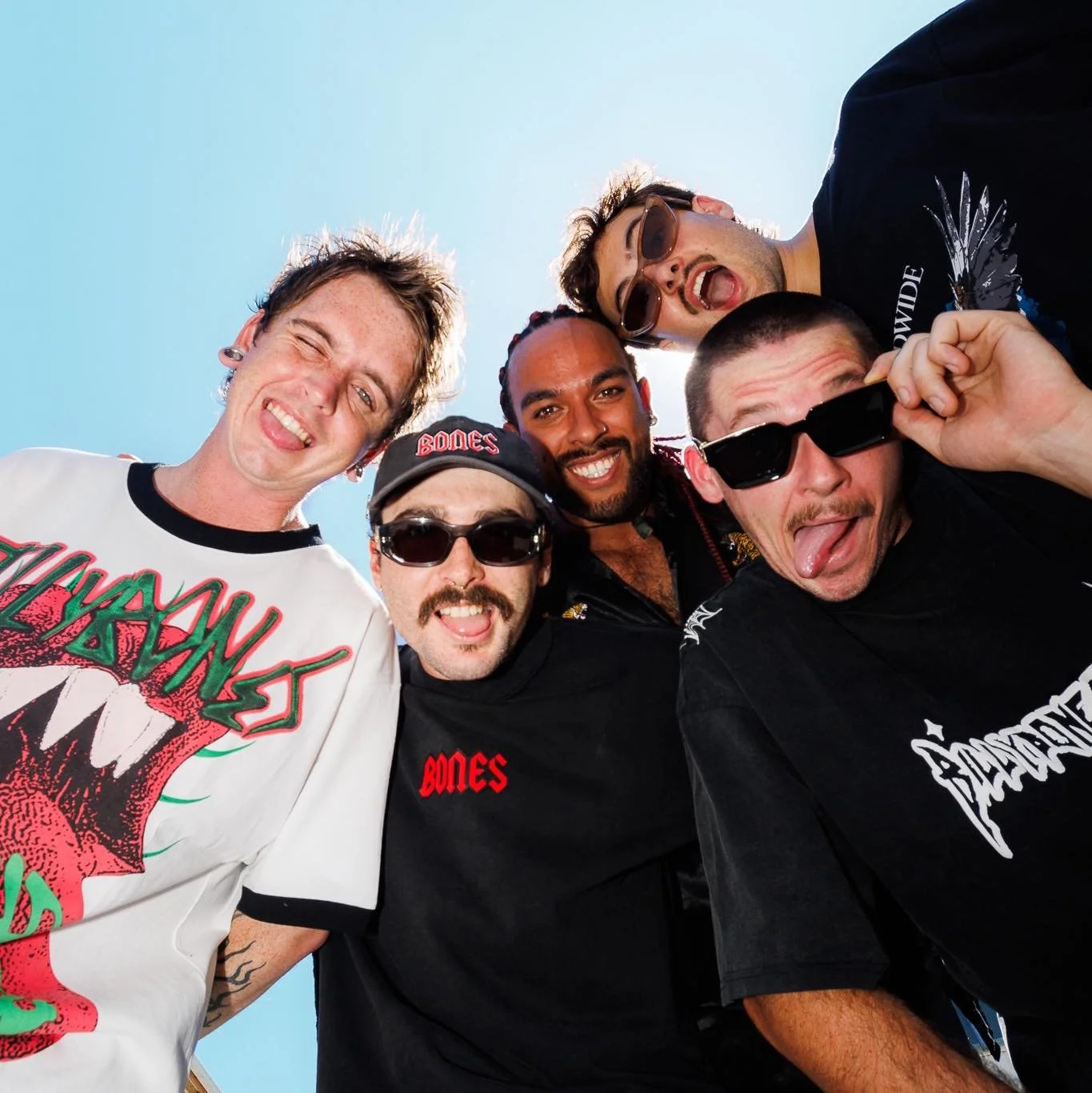 Group of six casually dressed men taking a selfie outdoors, smiling and making silly faces against a clear blue sky.