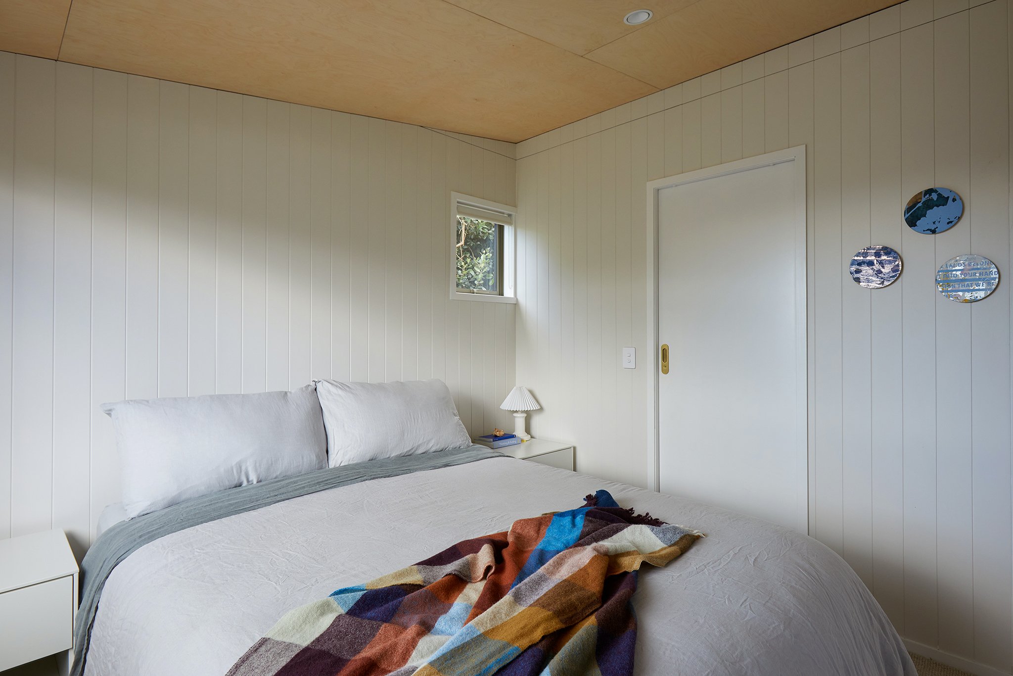 A bedroom with a bed, white bedding and a colorful blanket, a nightstand with a lamp, small window on the wall, and three decorative round mirrors on the wall.