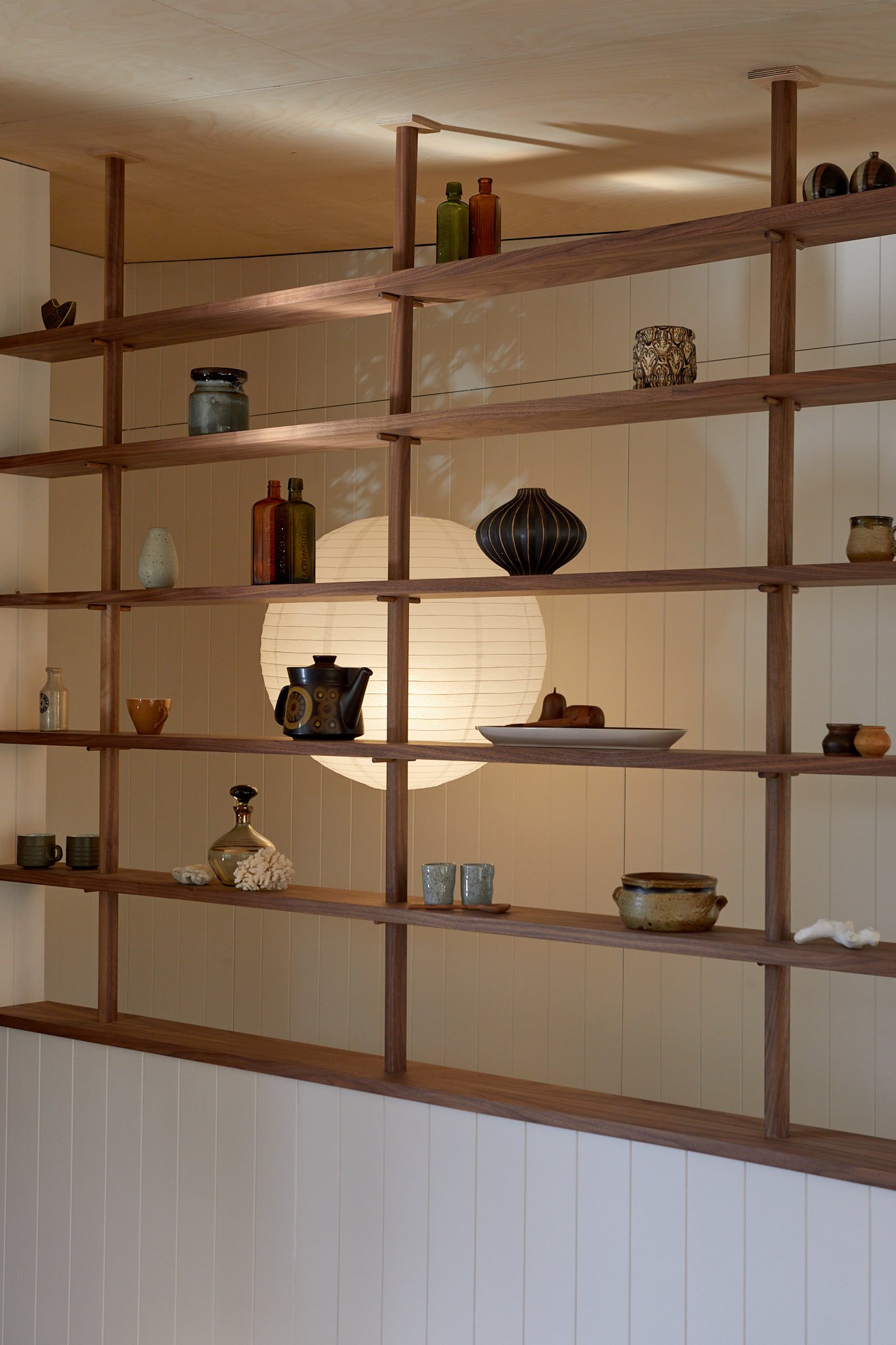 Wooden bookshelf with decorative vases, jars, cups, and a lamp, illuminated by a large paper lantern behind it.