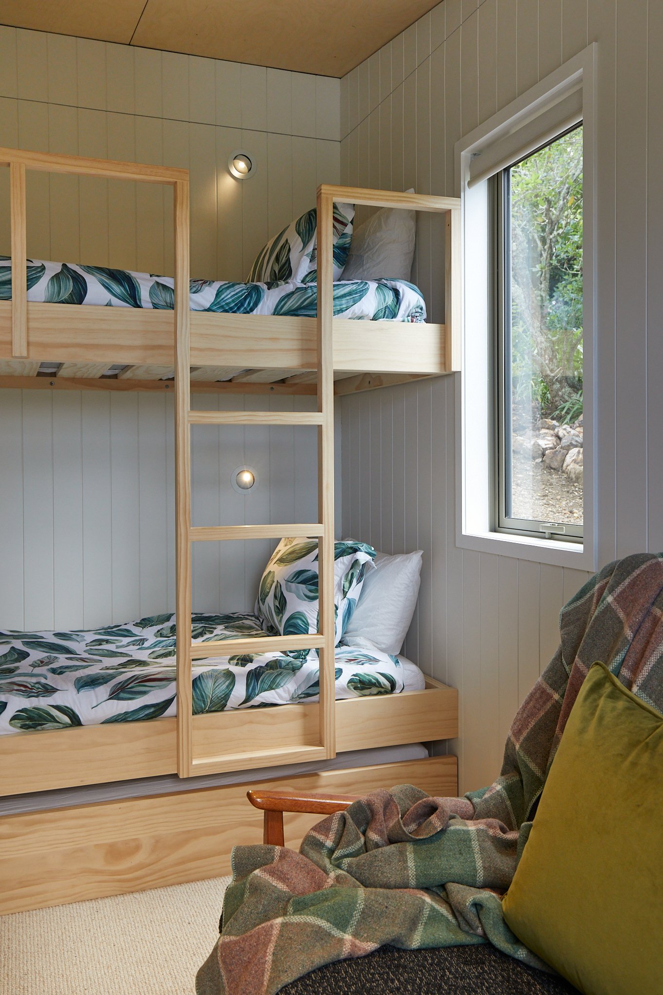 A cozy room with wooden bunk beds, featuring tropical leaf patterned bedding, a window showing outdoor greenery, and a plaid blanket draped over an armchair with a moss green pillow.