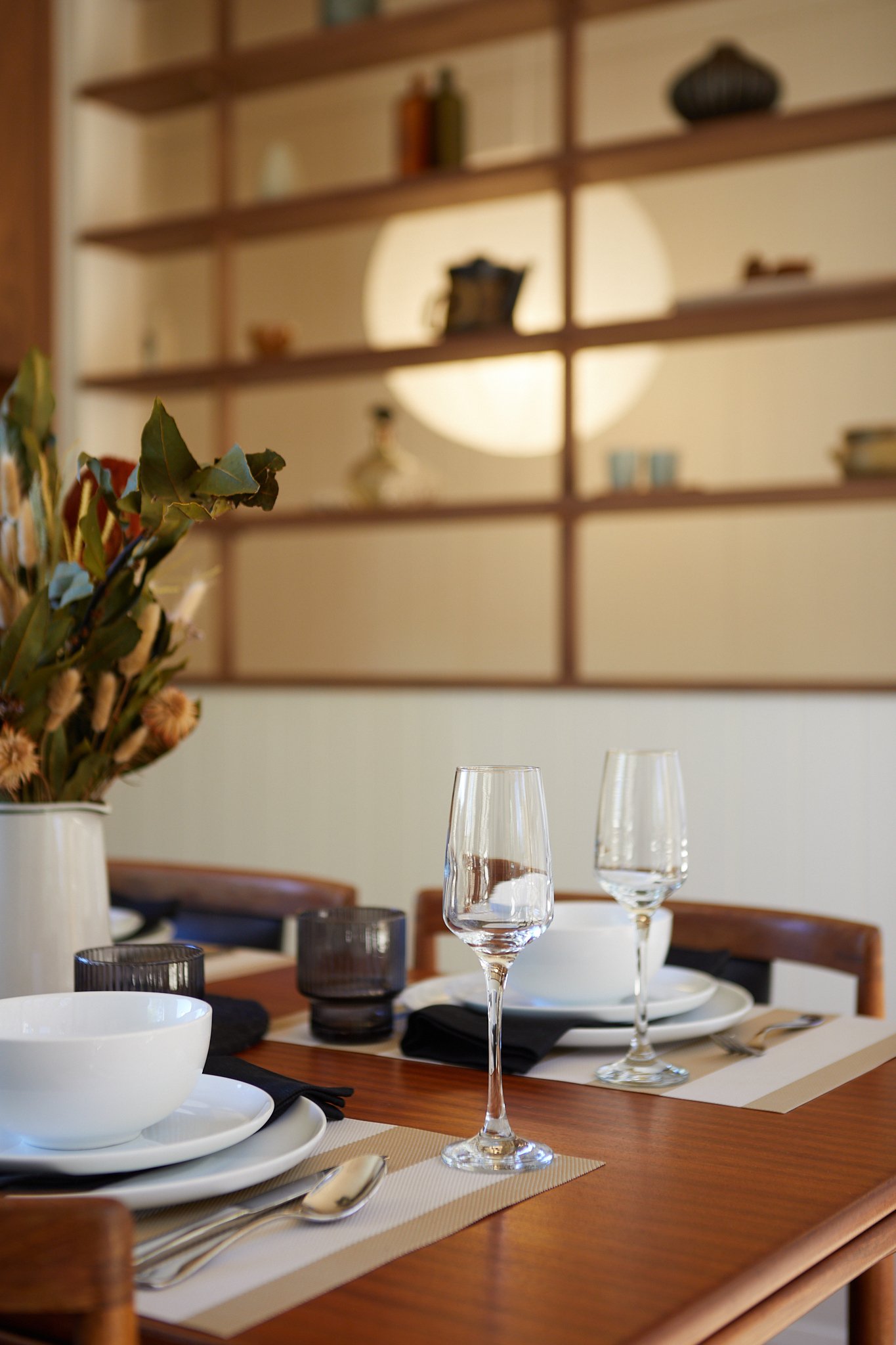 A neatly set dining table with white plates, wine glasses, cutlery, black napkins, and a vase with dried flowers in a cozy dining room.