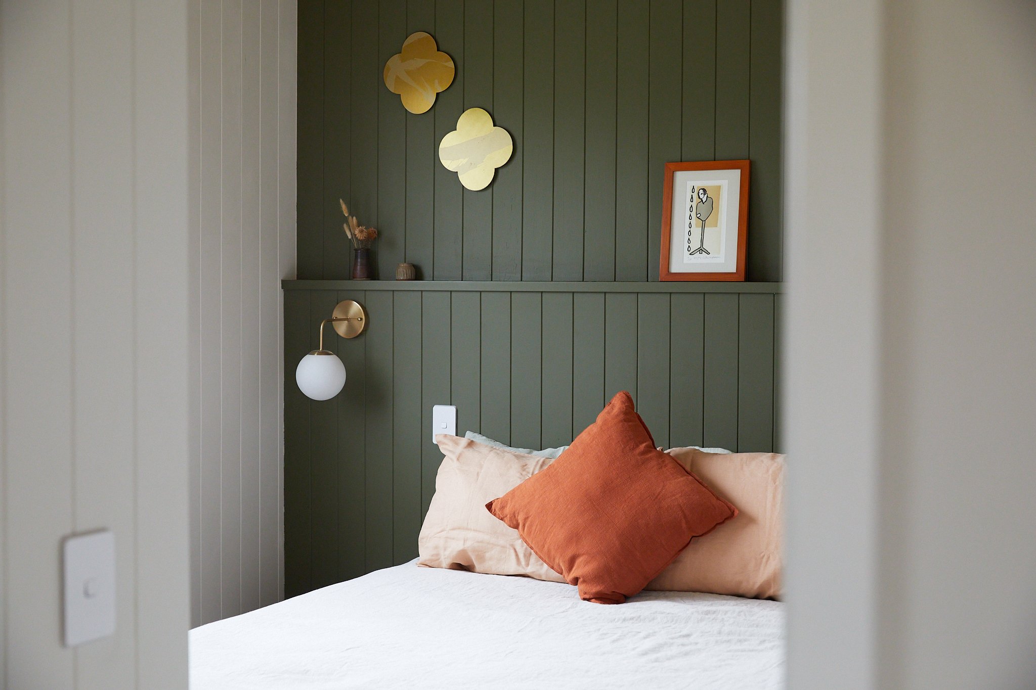 A bedroom with a bed featuring beige and orange pillows, a dark green paneled accent wall with three gold-shaped mirrors, a framed art print, and a wall-mounted lamp with a white globe shade.