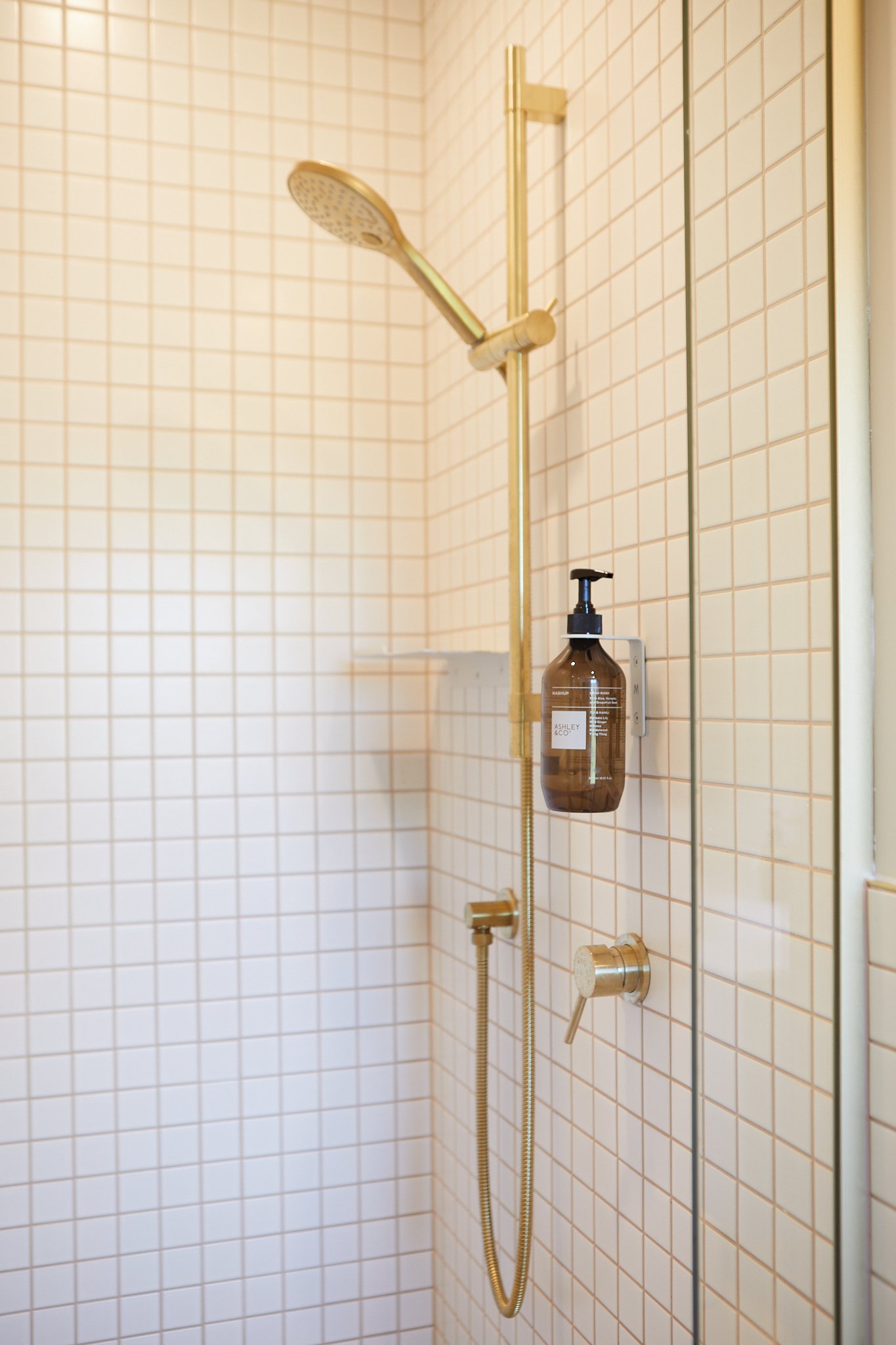 Close-up of a shower with gold fixtures, a hand-held showerhead, a wall-mounted soap dispenser, and a small angle of tiled wall.