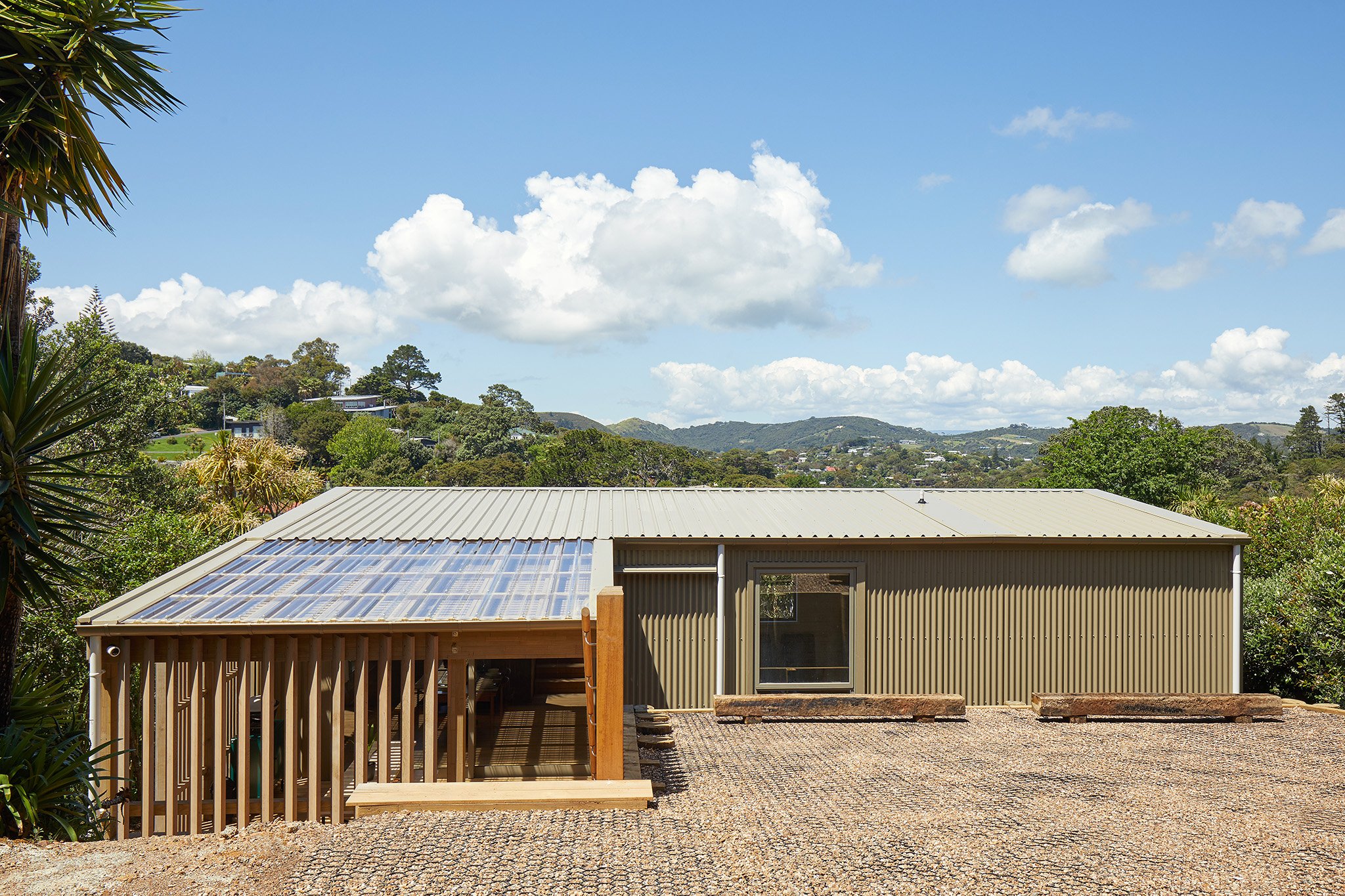 Modern house with metal roof, partially covered in solar panels, surrounded by trees and hills under a partly cloudy sky.