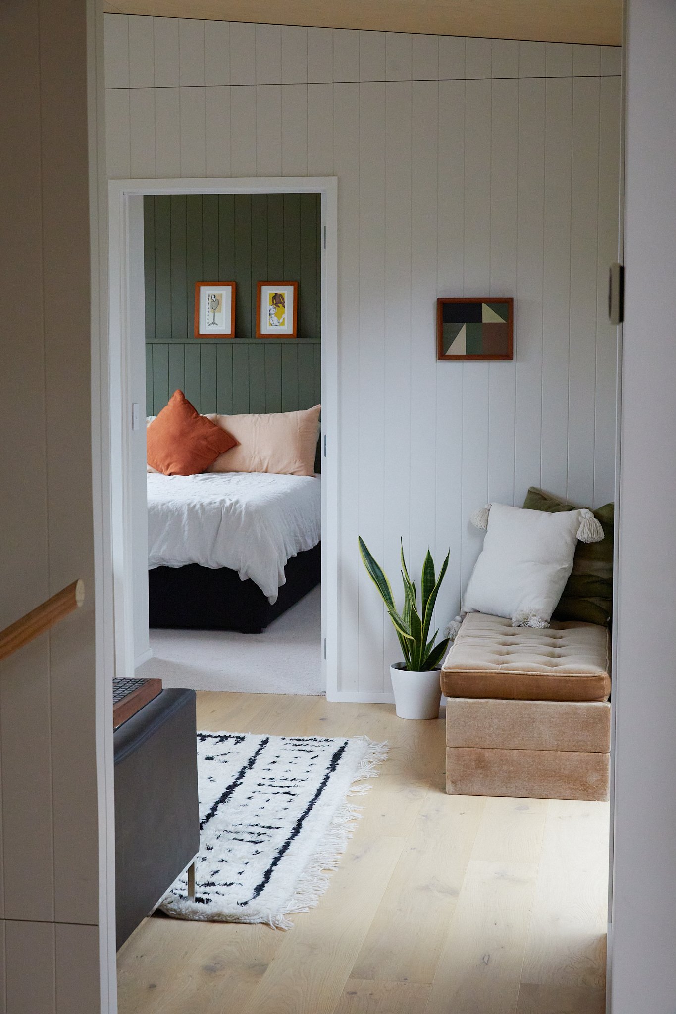 Interior view of a home showing a hallway with a small beige bench with a white pillow, a potted plant, and an abstract framed artwork on the wall; through an open doorway, a bedroom with a bed, throw pillows, and artwork on the wall can be seen.