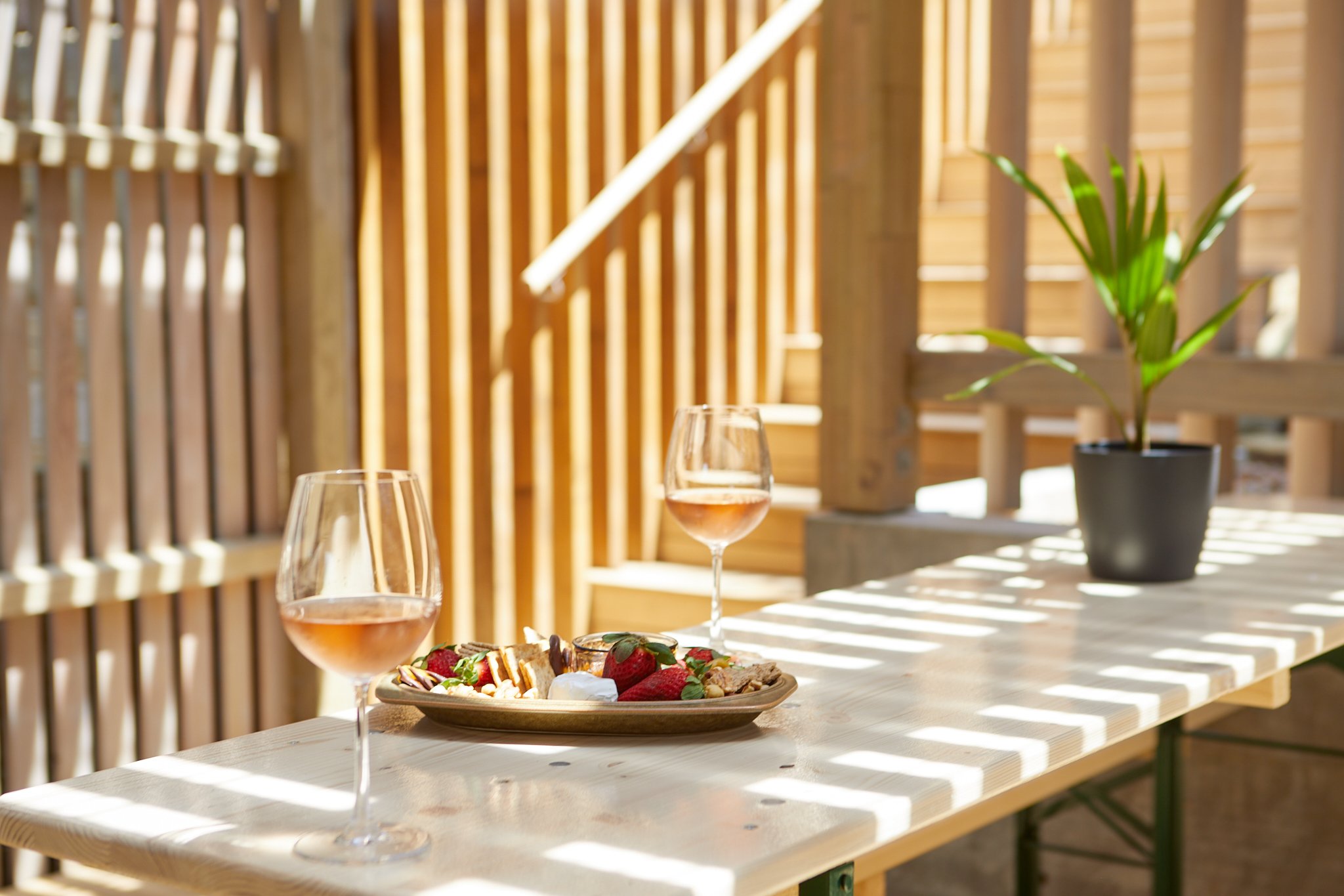 A wooden outdoor table with two glasses of rosé wine, a plate of strawberries, desserts, and a potted green plant. Sunlight filters through wooden slats casting striped shadows.