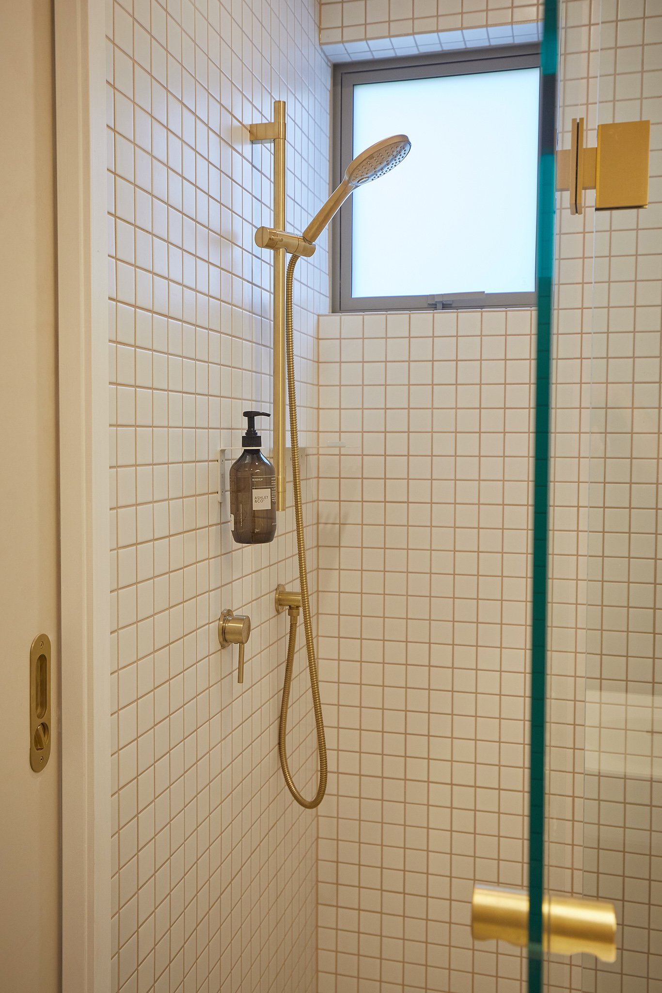 A small shower area with white tiled walls, a brass showerhead, a window, and a soap dispenser on the wall.