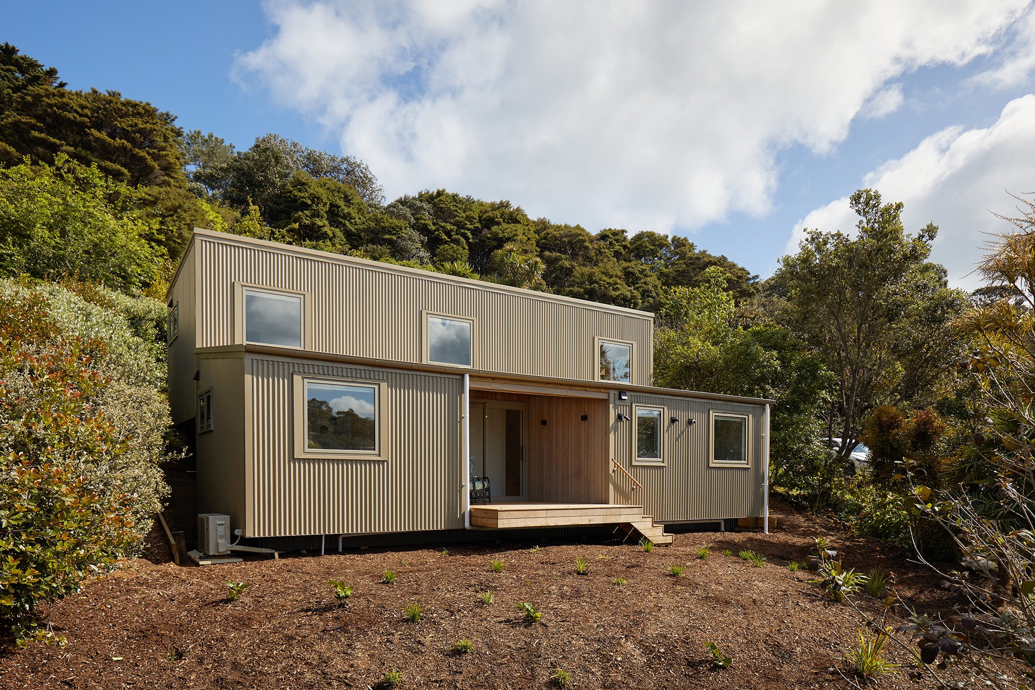Modern two-story house with vertical siding on a hillside, surrounded by trees and bushes, under a partly cloudy sky.