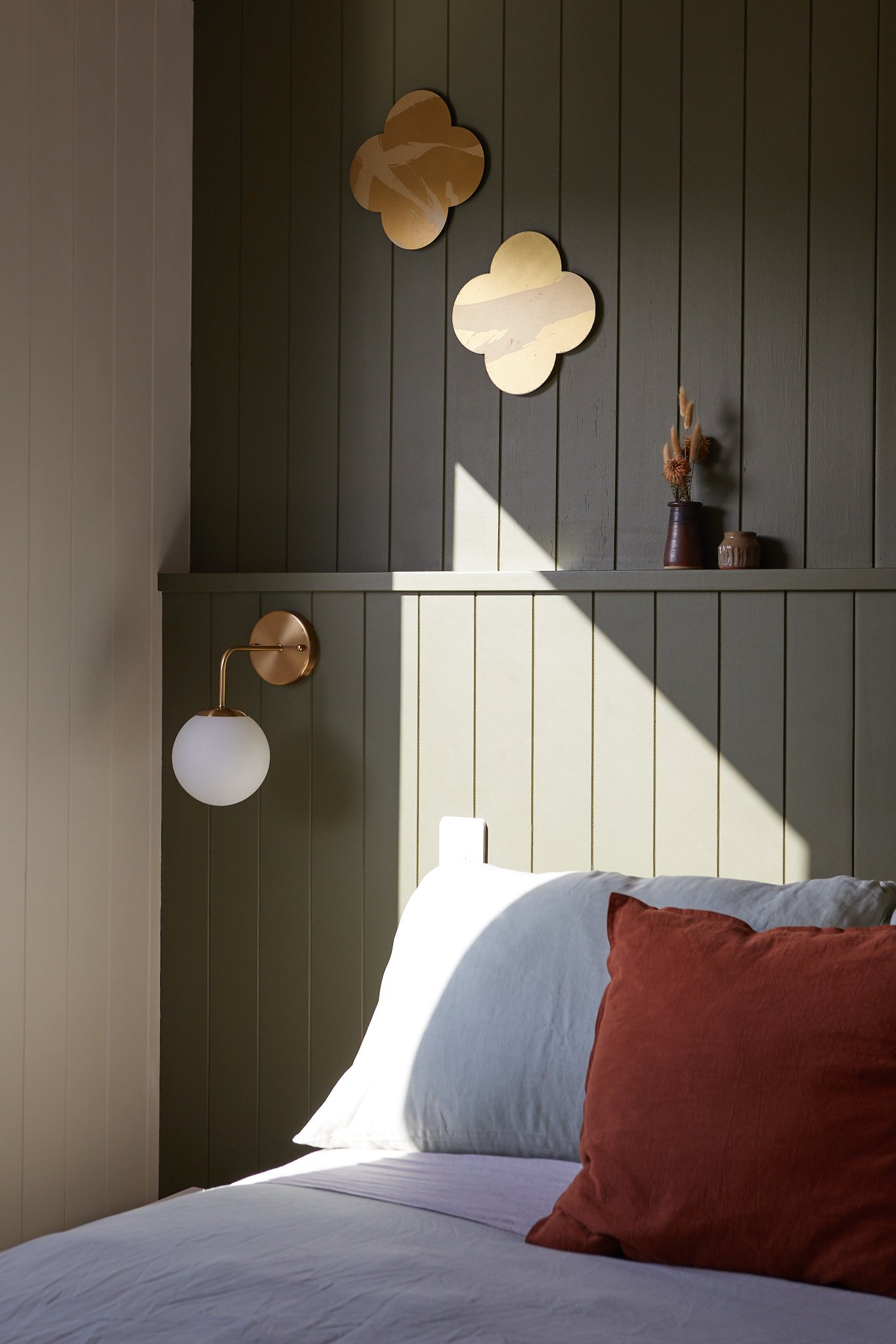 A bedroom with beige and gray bedding, olive green wooden paneled walls, decorative gold mirrors, and a wall-mounted white and gold sconce next to a small shelf with vases and dried flowers, illuminated by natural sunlight.