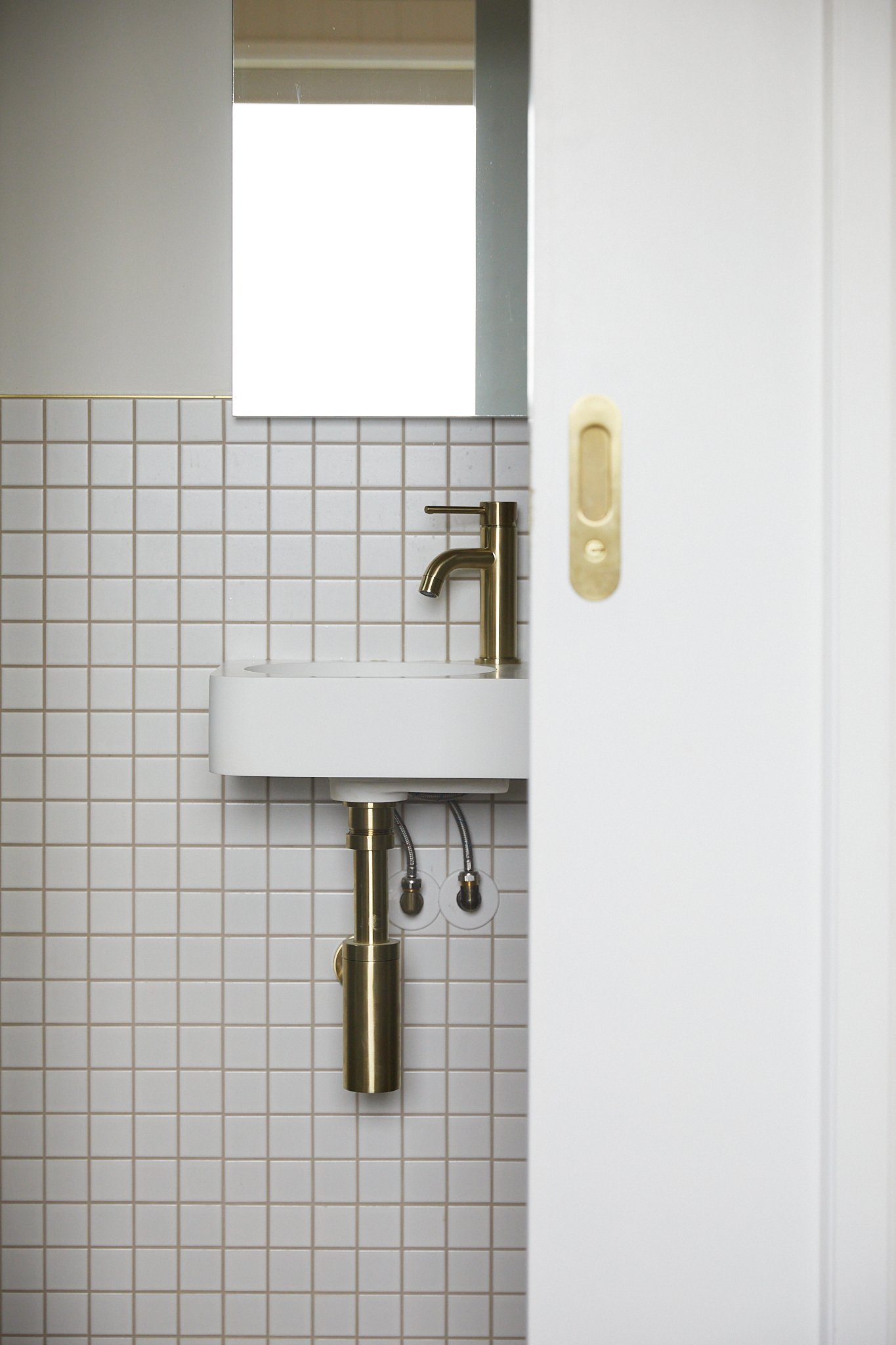 Small white bathroom sink with gold faucet, partially hidden behind a white sliding door, with beige square tile wall and mirror above.