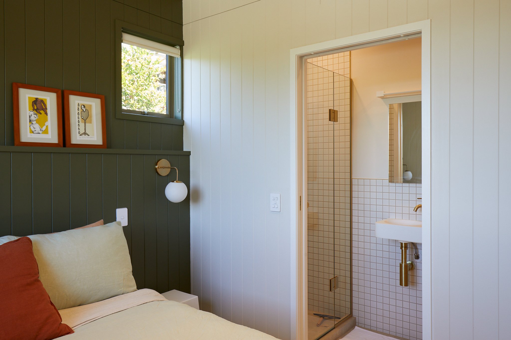 Bedroom with green and white paneled walls, and a view into a tiled bathroom with a glass shower door and white sink.