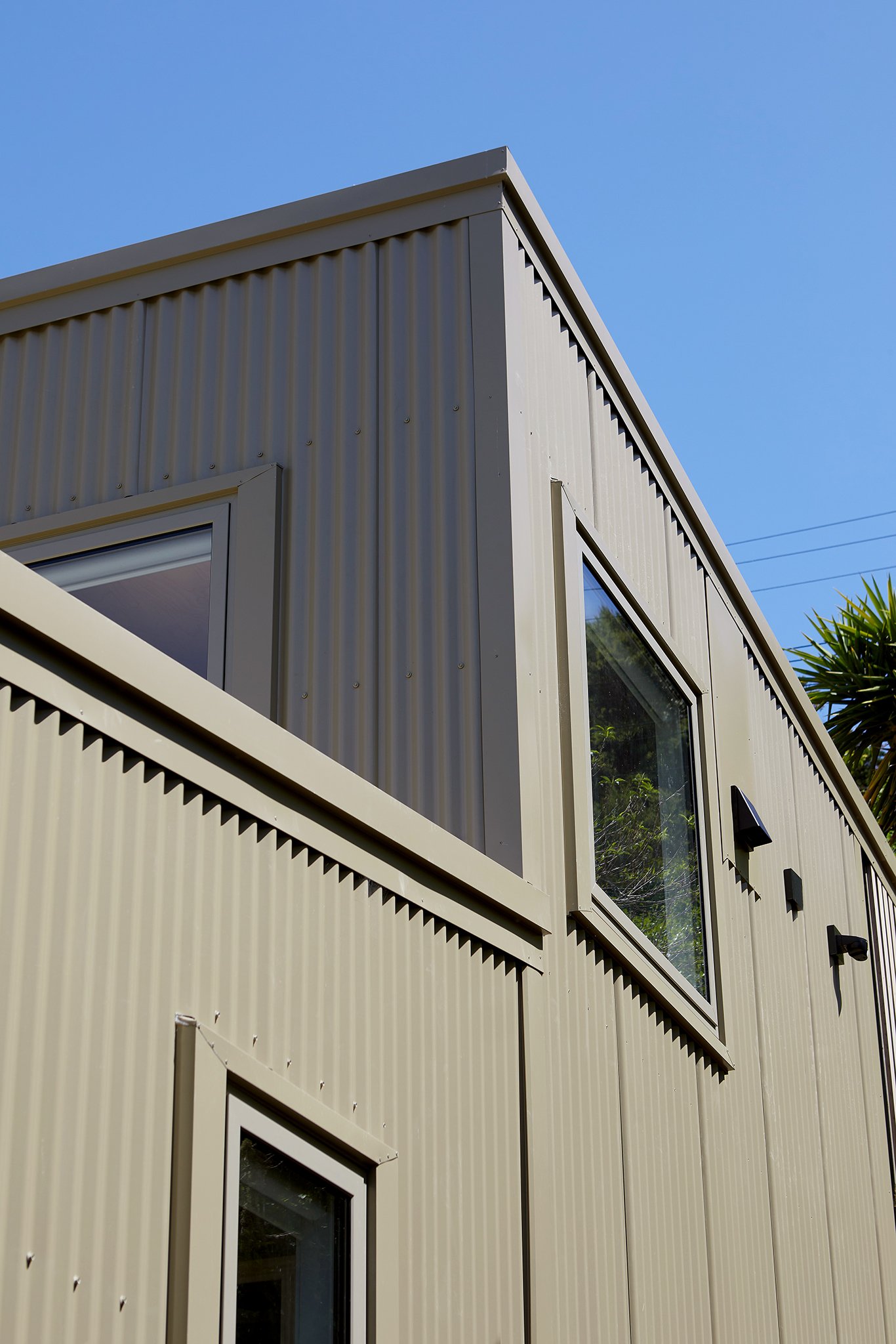 Close-up view of a beige metal building with vertical siding, multiple windows, and black exterior lights against a clear blue sky.