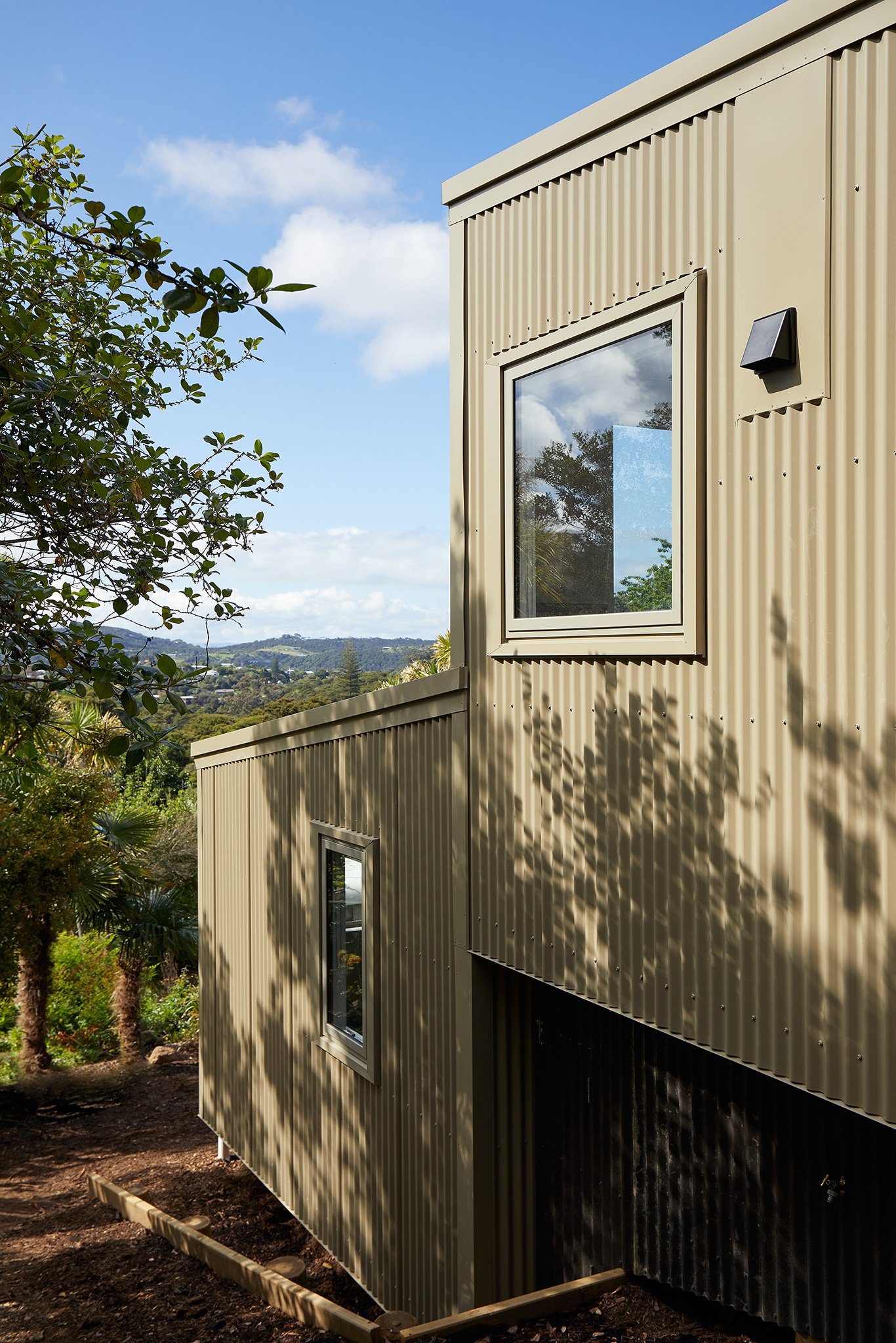 Modern beige metal building with rectangular windows, surrounded by trees and greenery, casting shadows on the exterior.