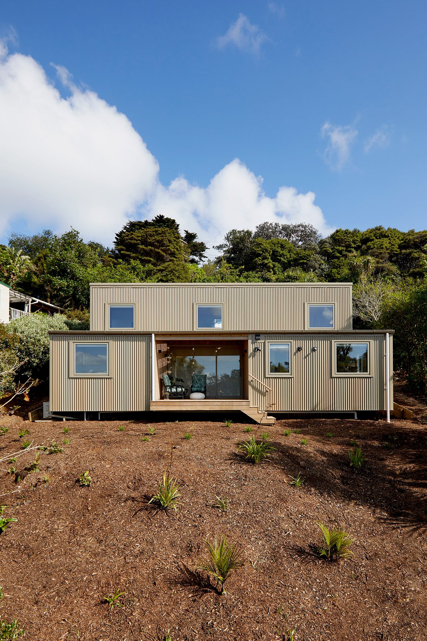 Modern beige house with metal siding and multiple windows, situated on a freshly landscaped yard with small plants, against a backdrop of green trees and a bright blue sky with scattered clouds.