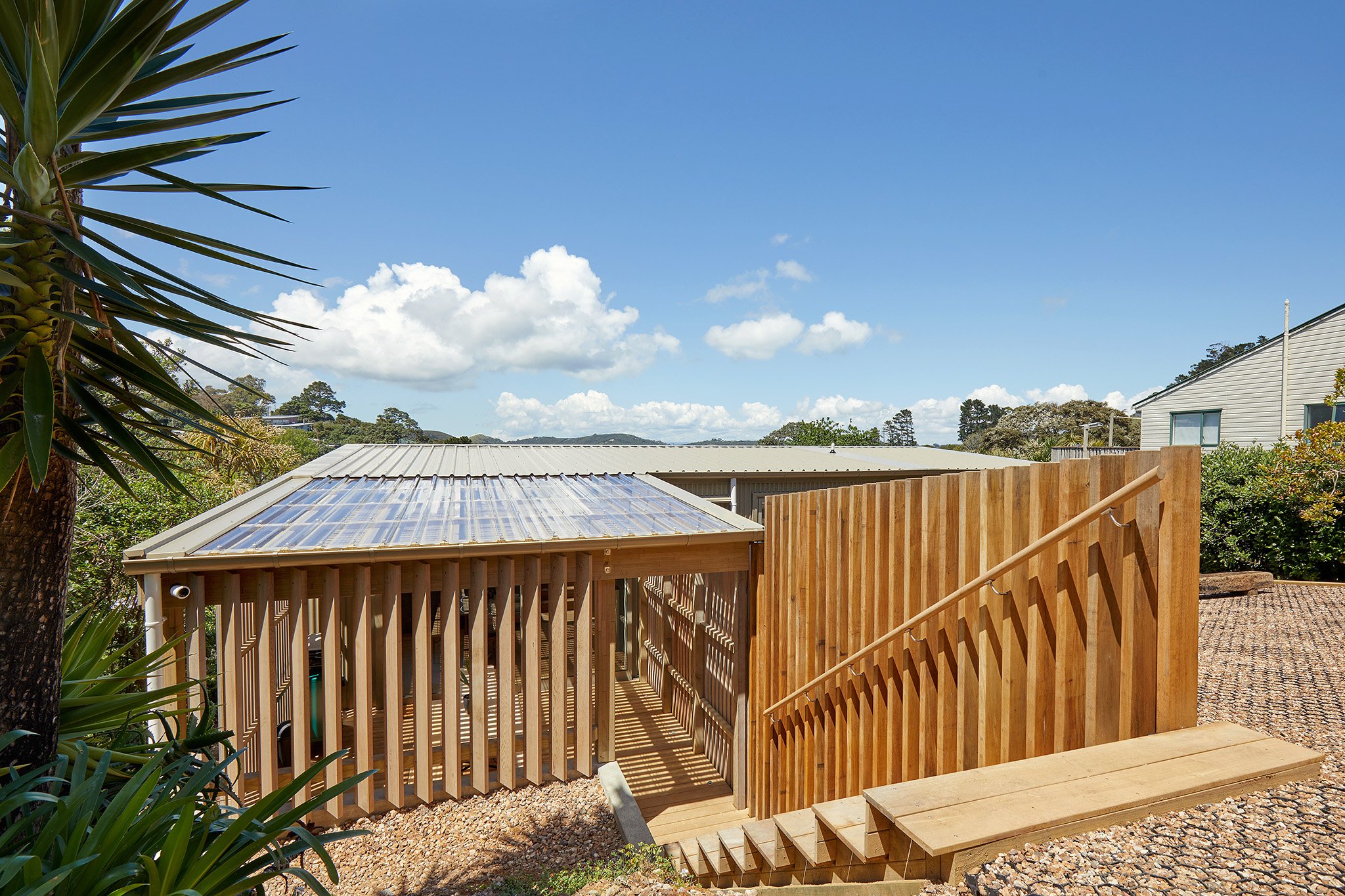 Modern house with wooden fencing, stairs, and a gravel yard under a blue sky with clouds.