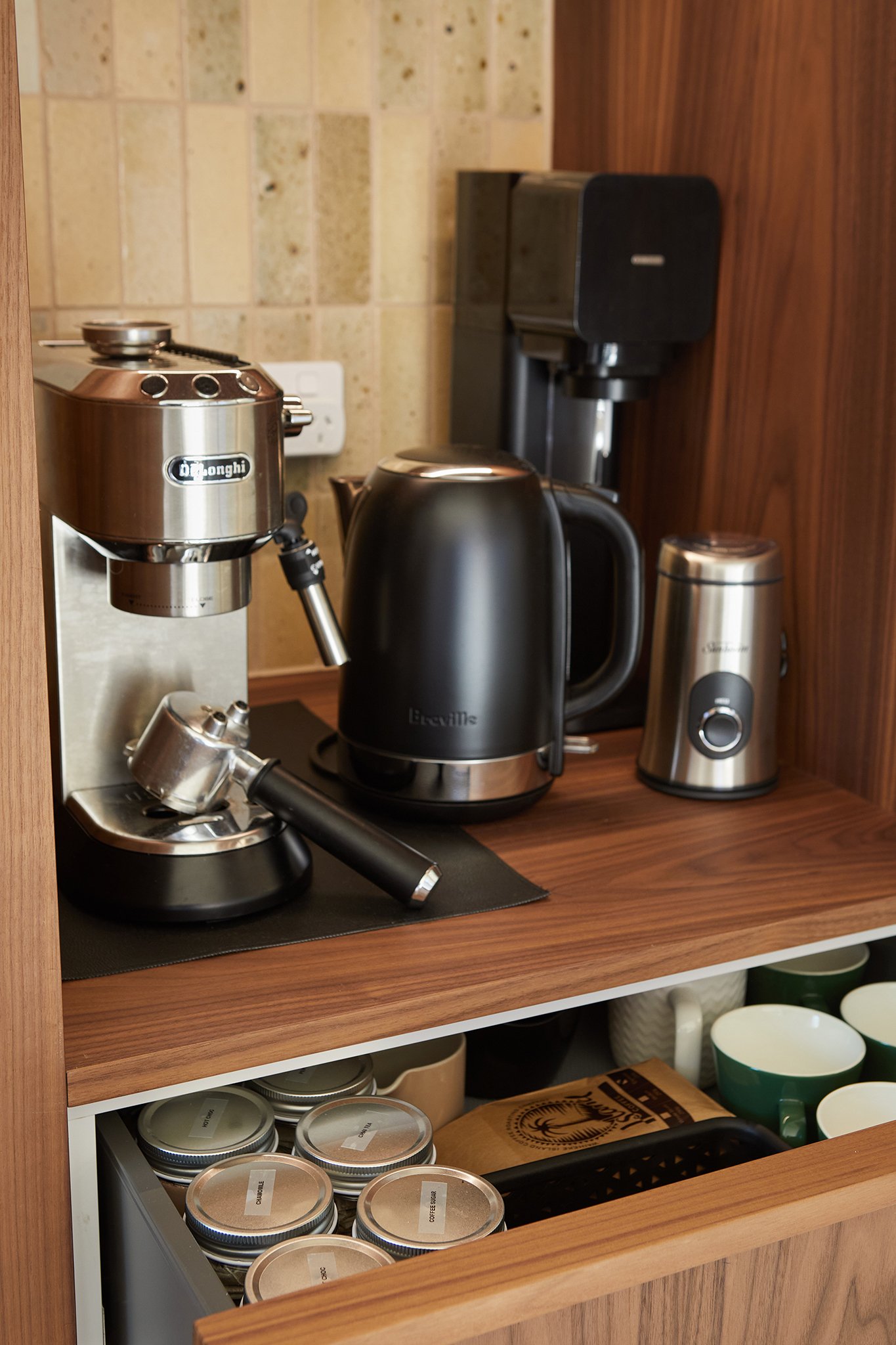 Coffee station with an espresso machine, electric kettle, coffee grinder, and coffee maker on a wooden countertop, with jars, mugs, and a paper bag of coffee below.