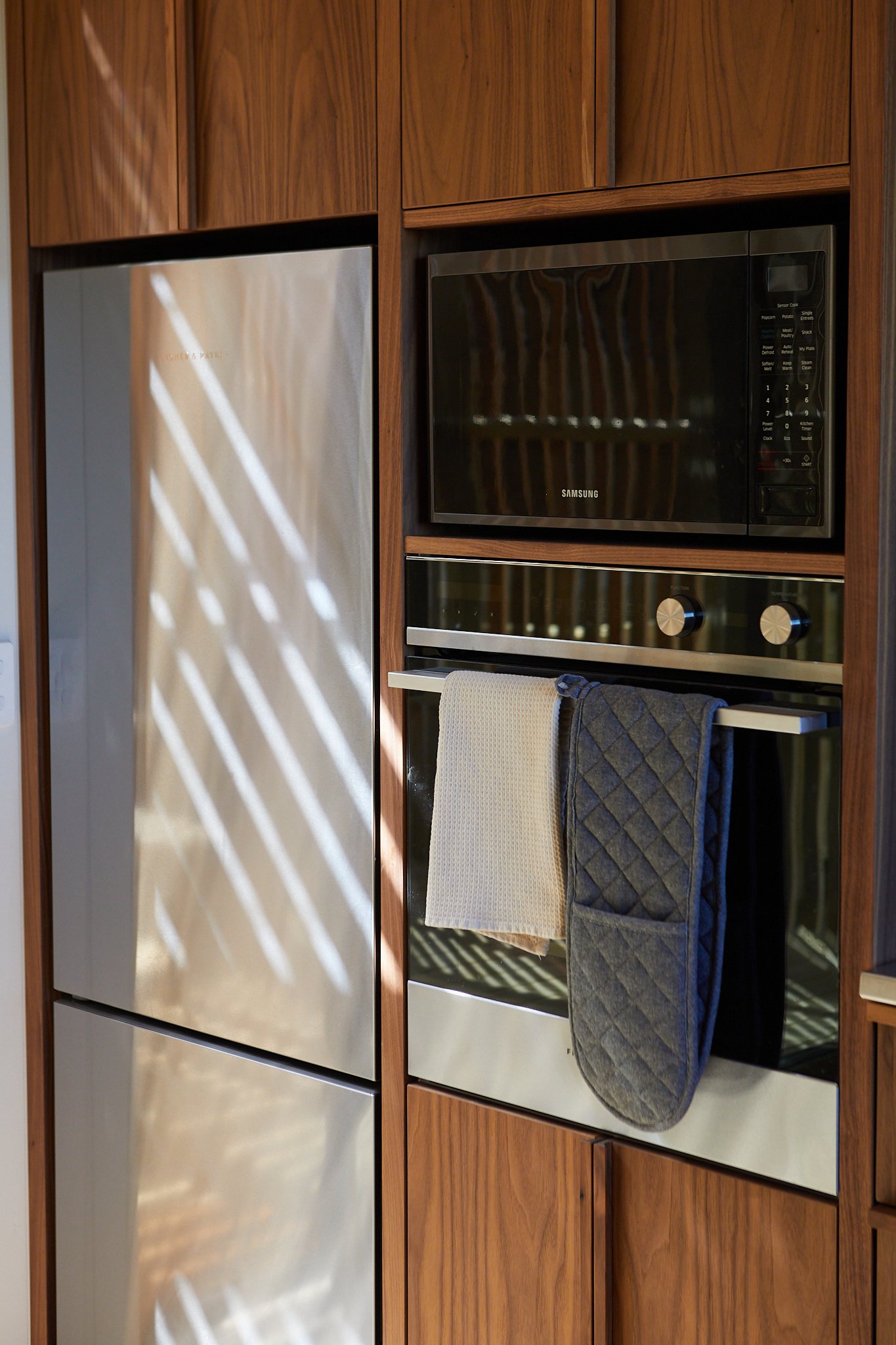 Kitchen with wooden cabinetry, a microwave oven, a built-in oven with oven mitts hanging on handle, and a stainless steel refrigerator.