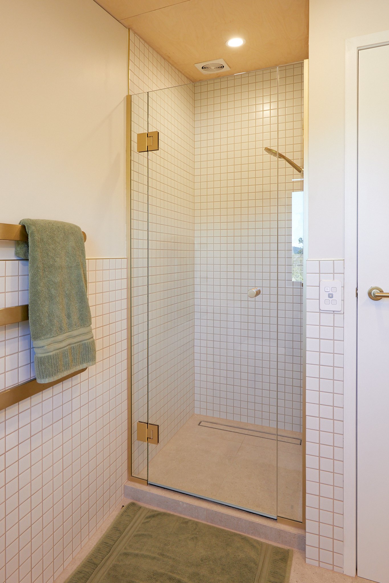 Modern bathroom with a walk-in shower enclosed by glass, with a beige tile floor, small beige tiles on the walls, a towel rack with a green towel, and a white door.