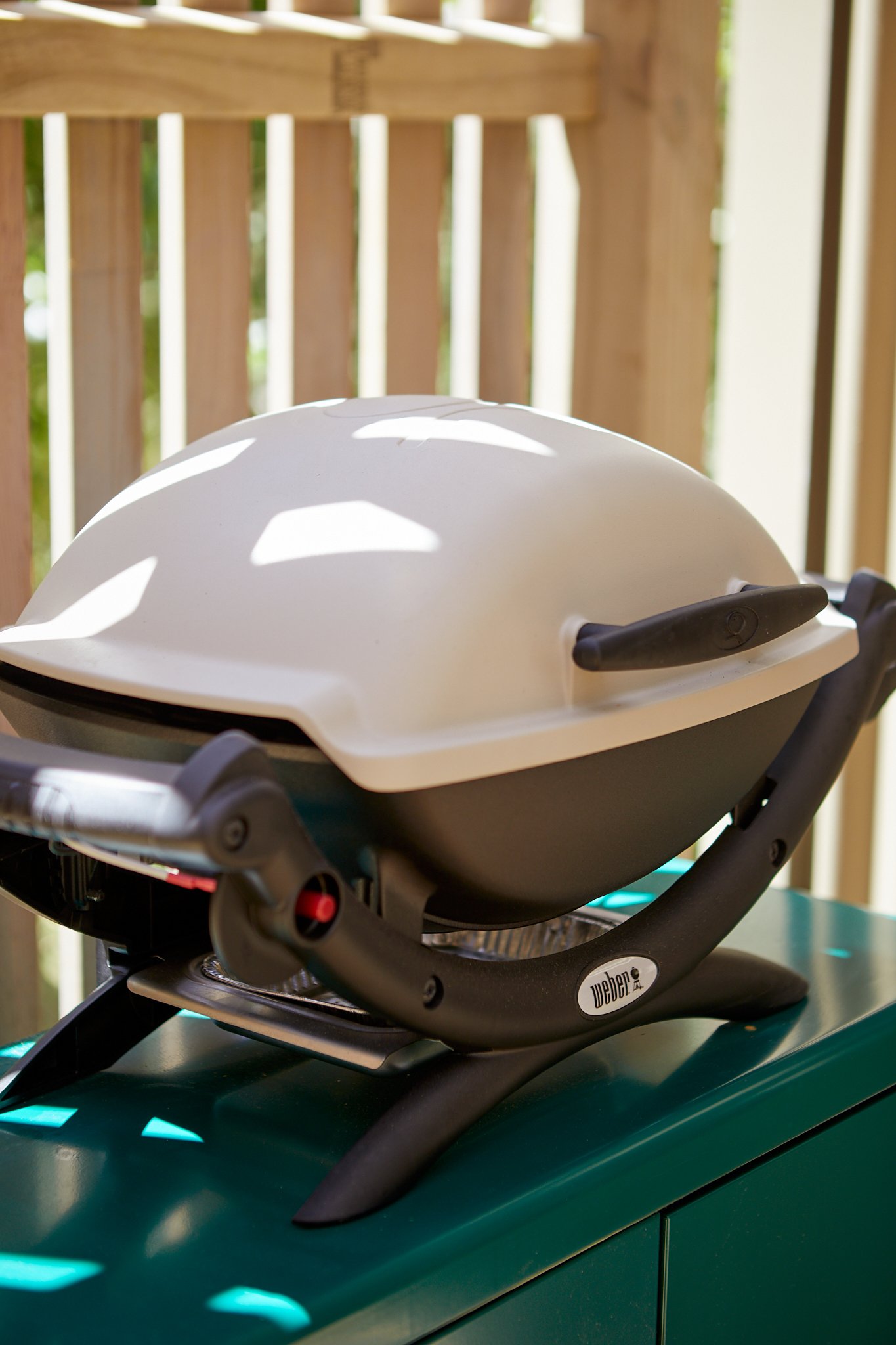 A small, round outdoor grill with a white lid placed on a teal-colored table or counter on a wooden deck, with a wooden railing in the background.