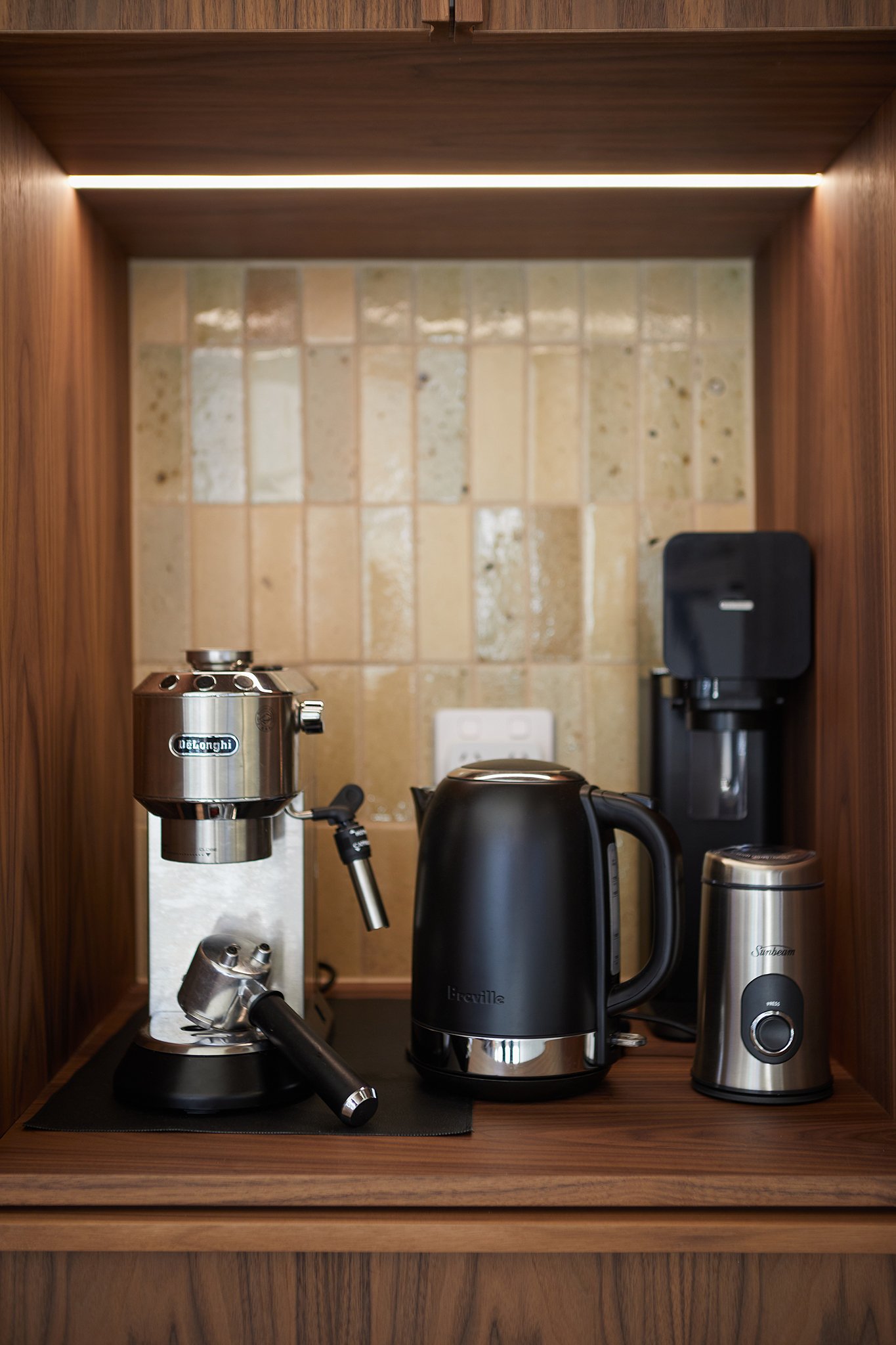 Coffee station with an espresso machine, electric kettle, coffee grinder, and coffee maker in a wooden cabinet under a strip of lighting.