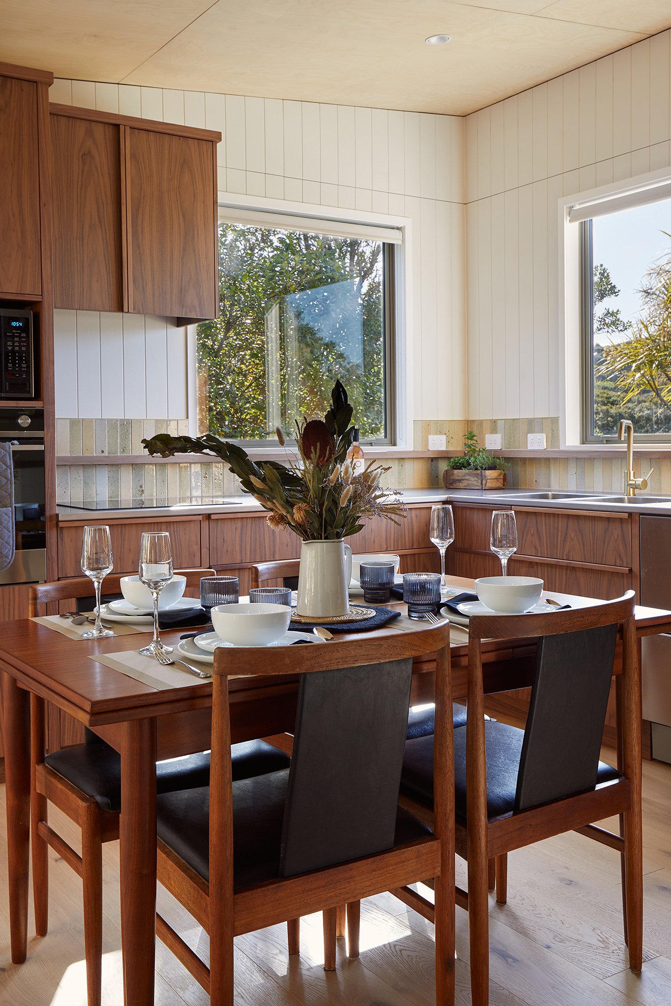 A dining table set for a meal in a bright kitchen with wooden cabinets, white panel walls, and large windows showing outdoor greenery.
