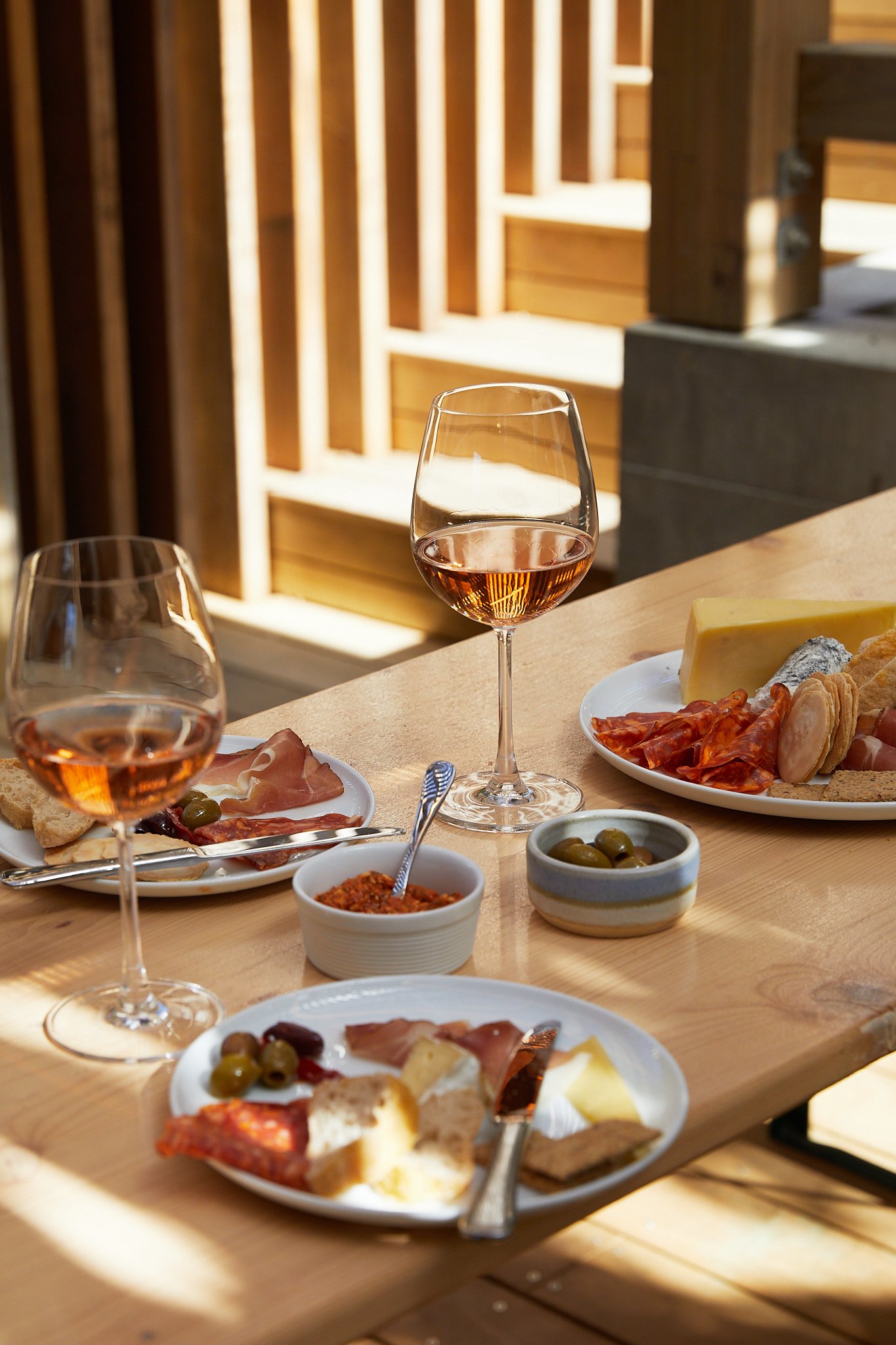 A wooden table set with two glasses of rosé wine, plates of assorted cheeses, meats, and crackers, small bowls of olives and spread, and a partially eaten cheese plate, in a sunny setting.