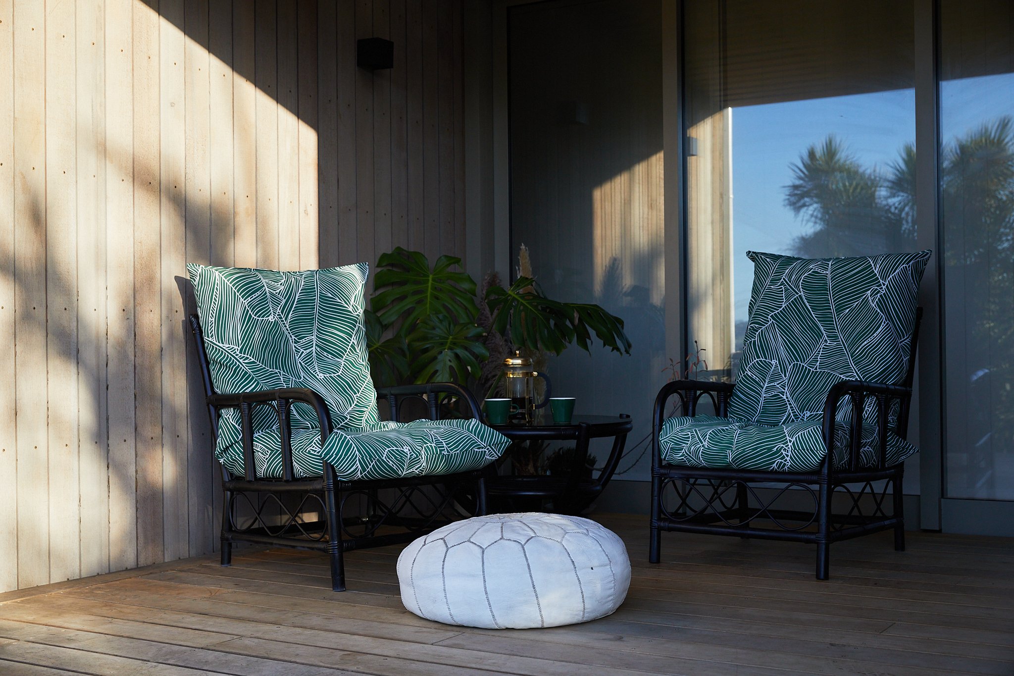 Outdoor patio with two black wicker chairs with large green leaf patterned cushions, a small black table with green cups and a glass pitcher, a white pouf, a large leafy plant, and a glass wall with a view of trees outside.