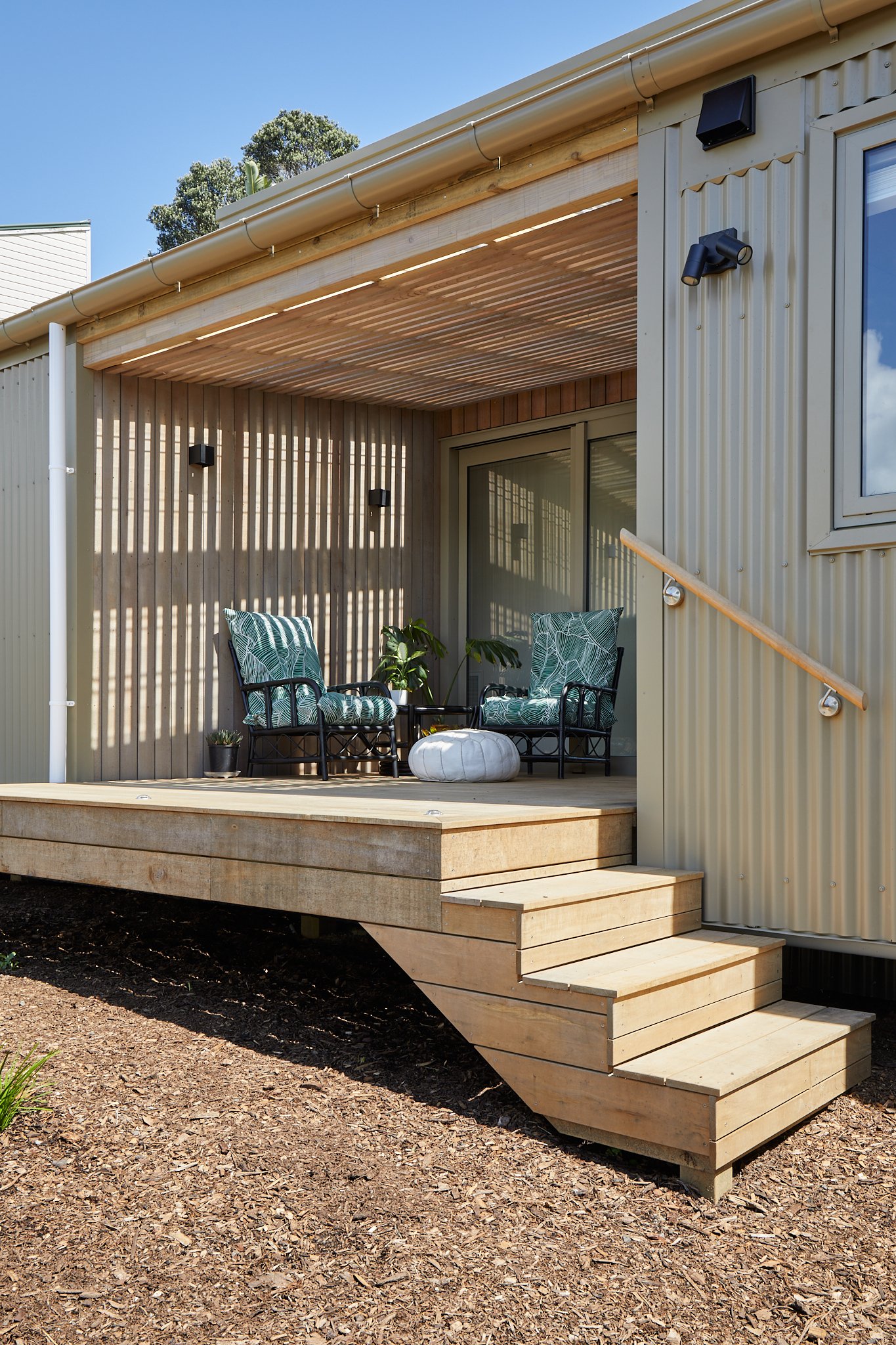 A small wooden deck with two chairs and a small table, attached to a modern tiny house with exterior wall lights and a sliding door, under a blue sky.