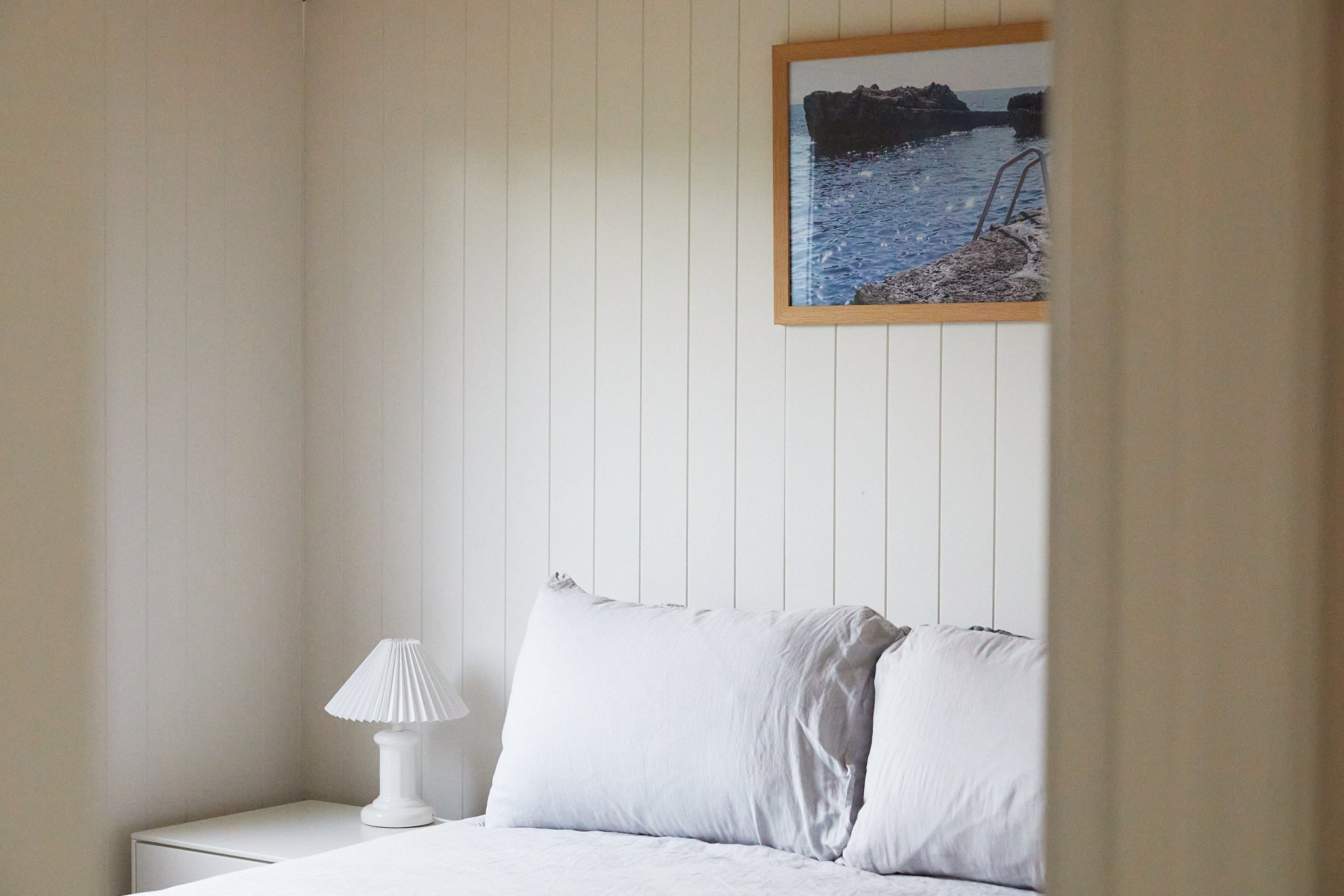A bedroom with white paneled walls, white bedding, a globe-shaped white lamp on a nightstand, and a framed photo of a rocky shoreline with water.