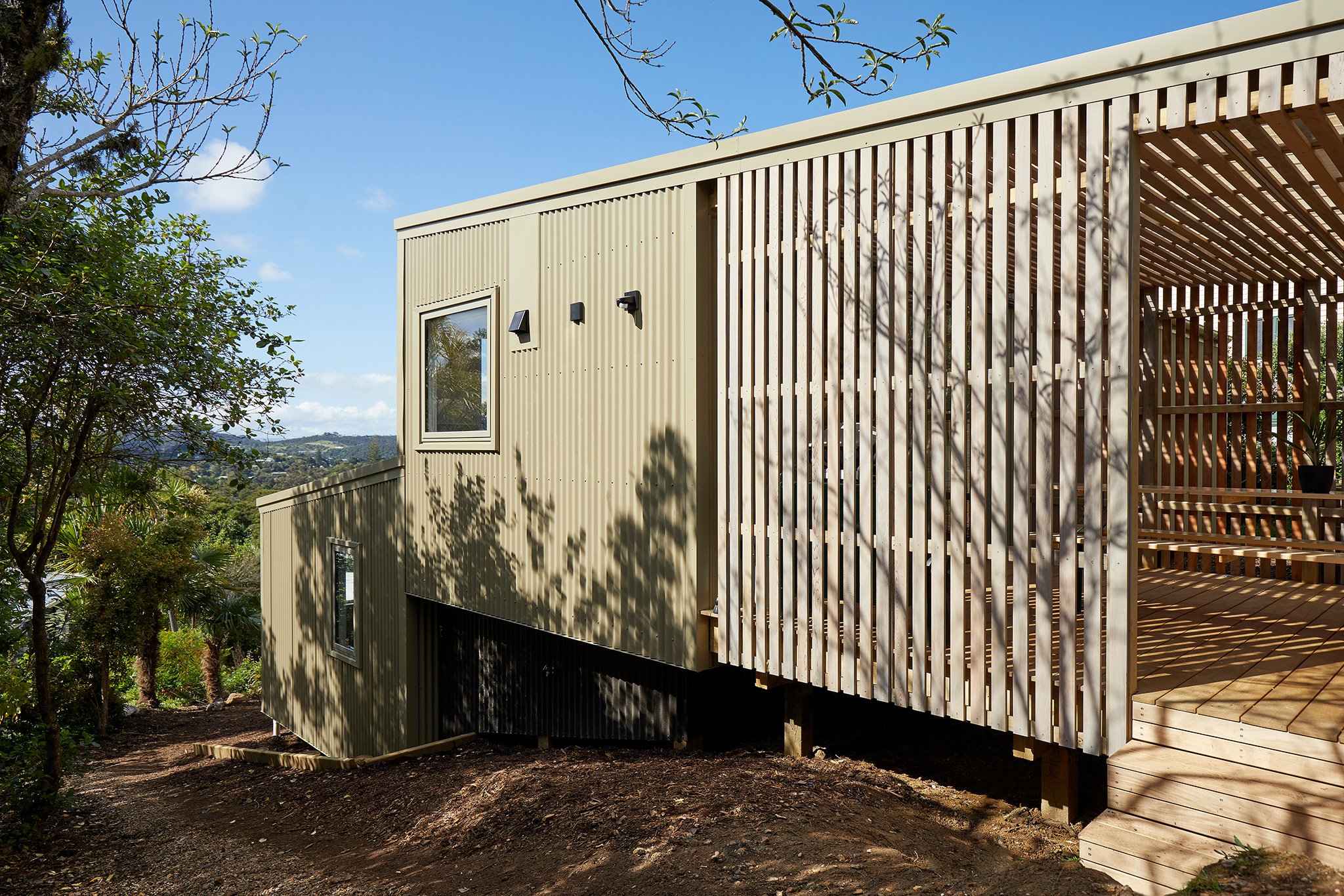 Modern house on a hillside with beige and wood exterior, surrounded by greenery and trees, under a blue sky.