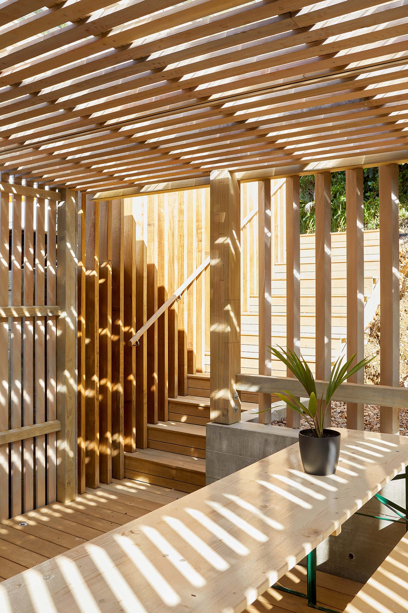 Interior view of a wooden patio with sunlight creating striped shadows on the ceiling, railing, and table, featuring a potted plant on the table.