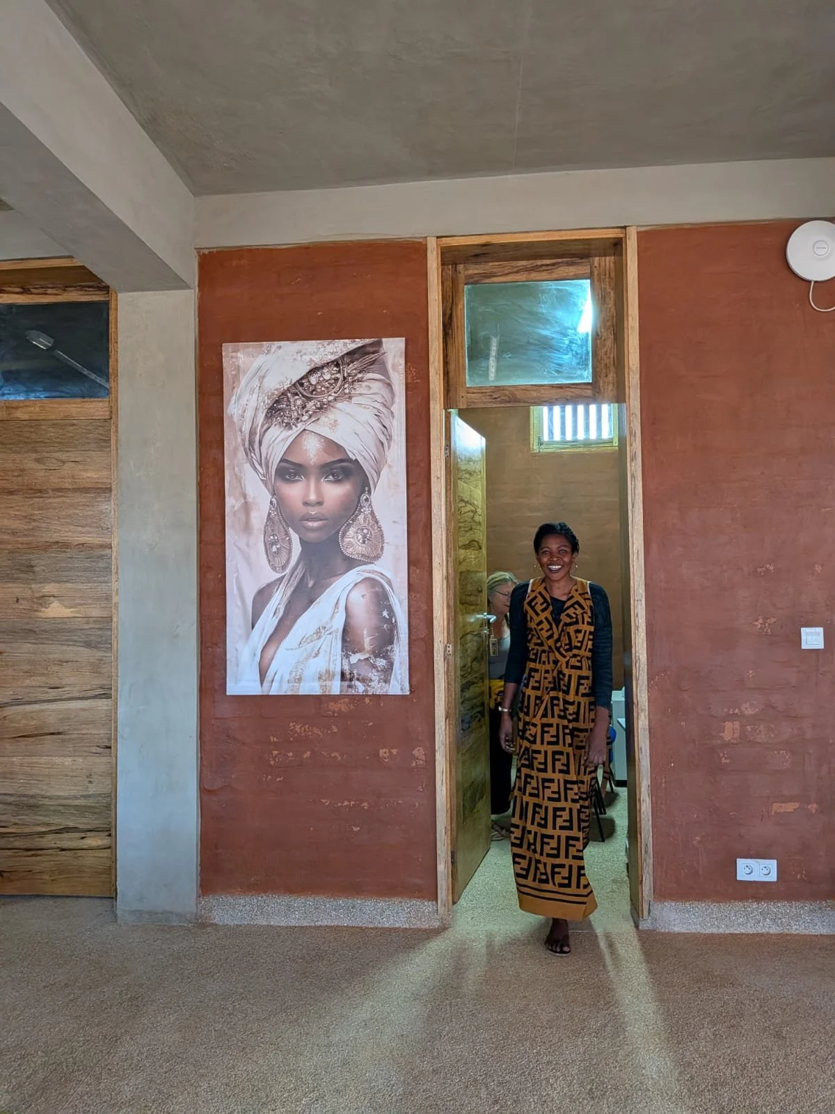 Mujer sonriendo caminando por una sala con paredes de color terracota, vestida con un vestido largo de estampado Fendi, y un póster grande de una mujer con un turbante y accesorios dorados en la pared.