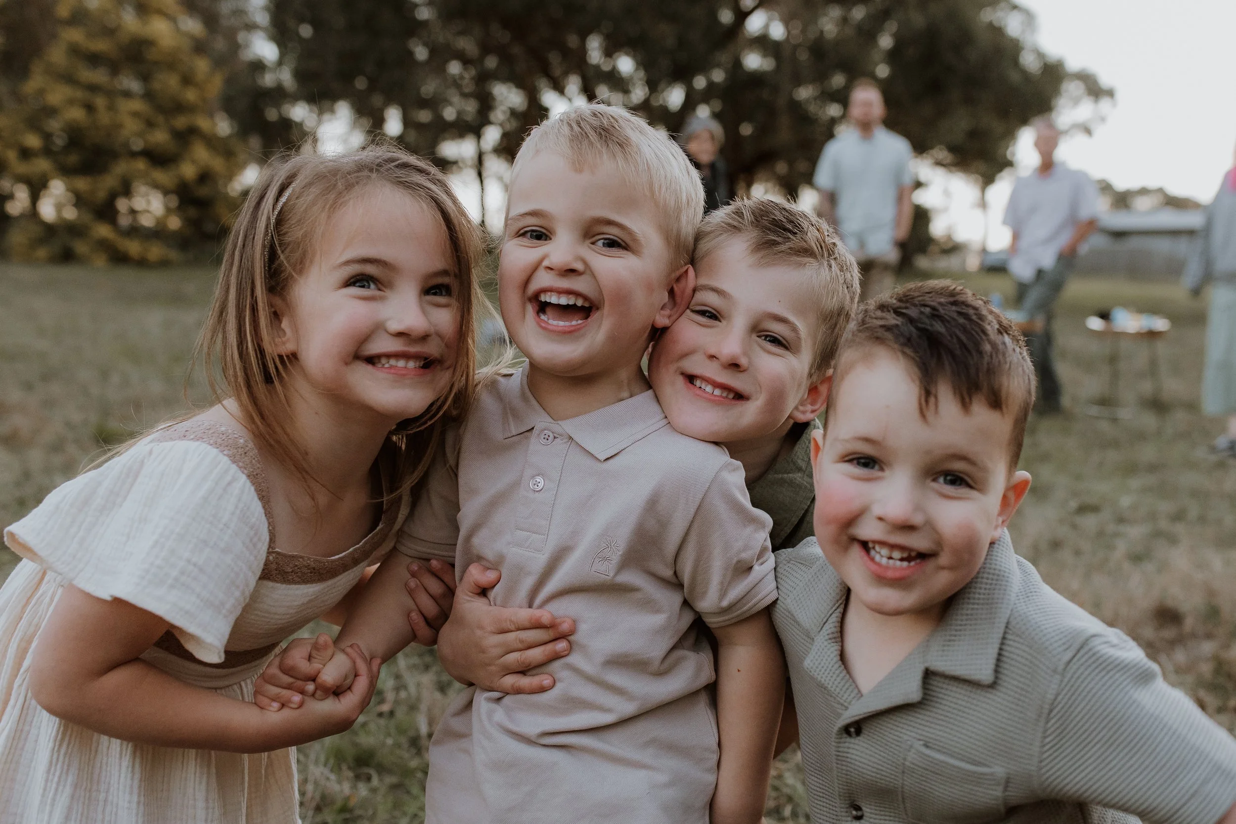 Group of smiling children hugging and leaning on each other outdoors in a park, with adults in the background and trees.