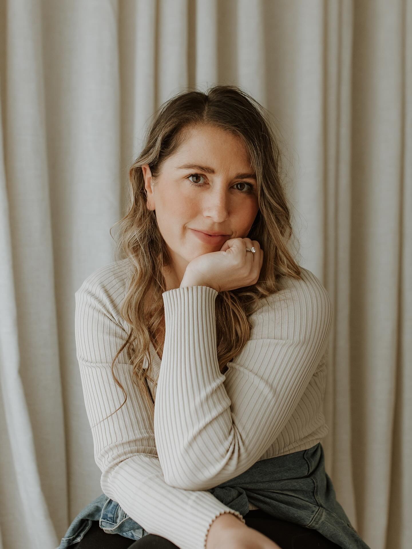 A woman with long wavy brown hair and light skin, sitting in front of beige curtains, wearing a cream-colored ribbed sweater and dark pants, resting her chin on her hand and looking at the camera.