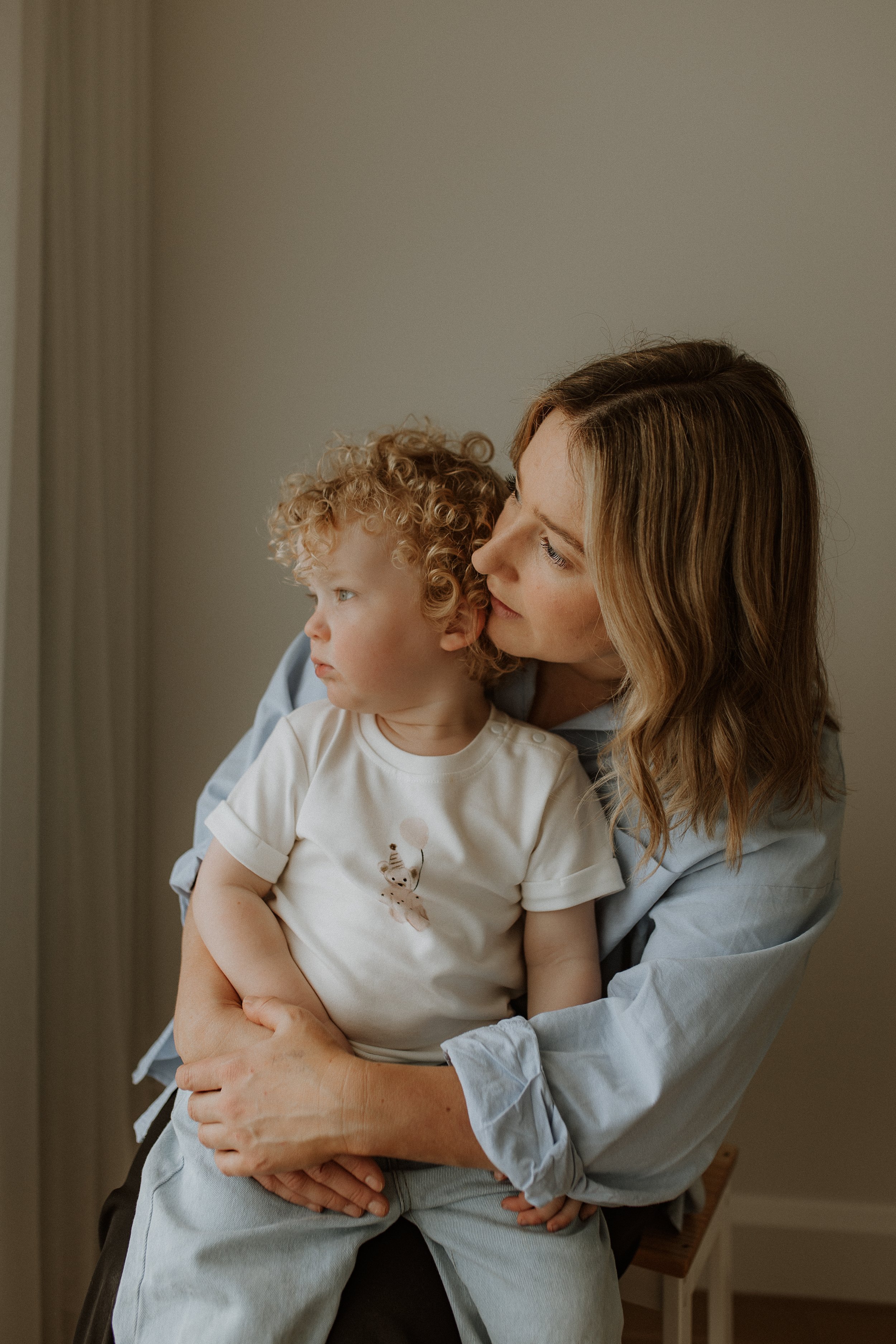 A woman with long, wavy hair holding a young boy with curly hair, both looking to the left, indoors against a neutral-colored wall.