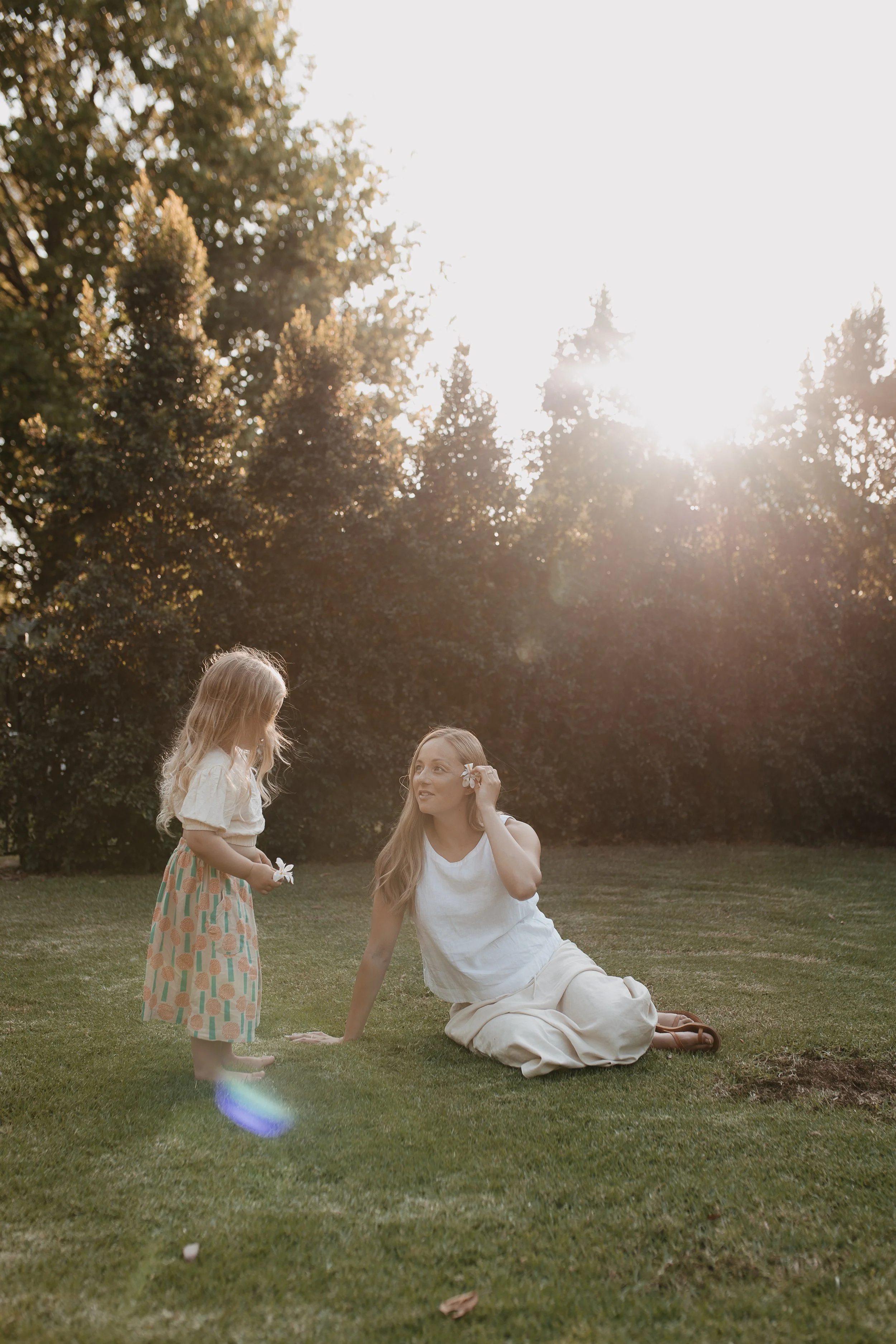 A woman with long hair sitting on the grass, wearing a white sleeveless top and beige pants, looking up at a young girl. The girl wears a patterned skirt and a light top, holding a small flower. They are outdoors in a park with tall trees and sunligh