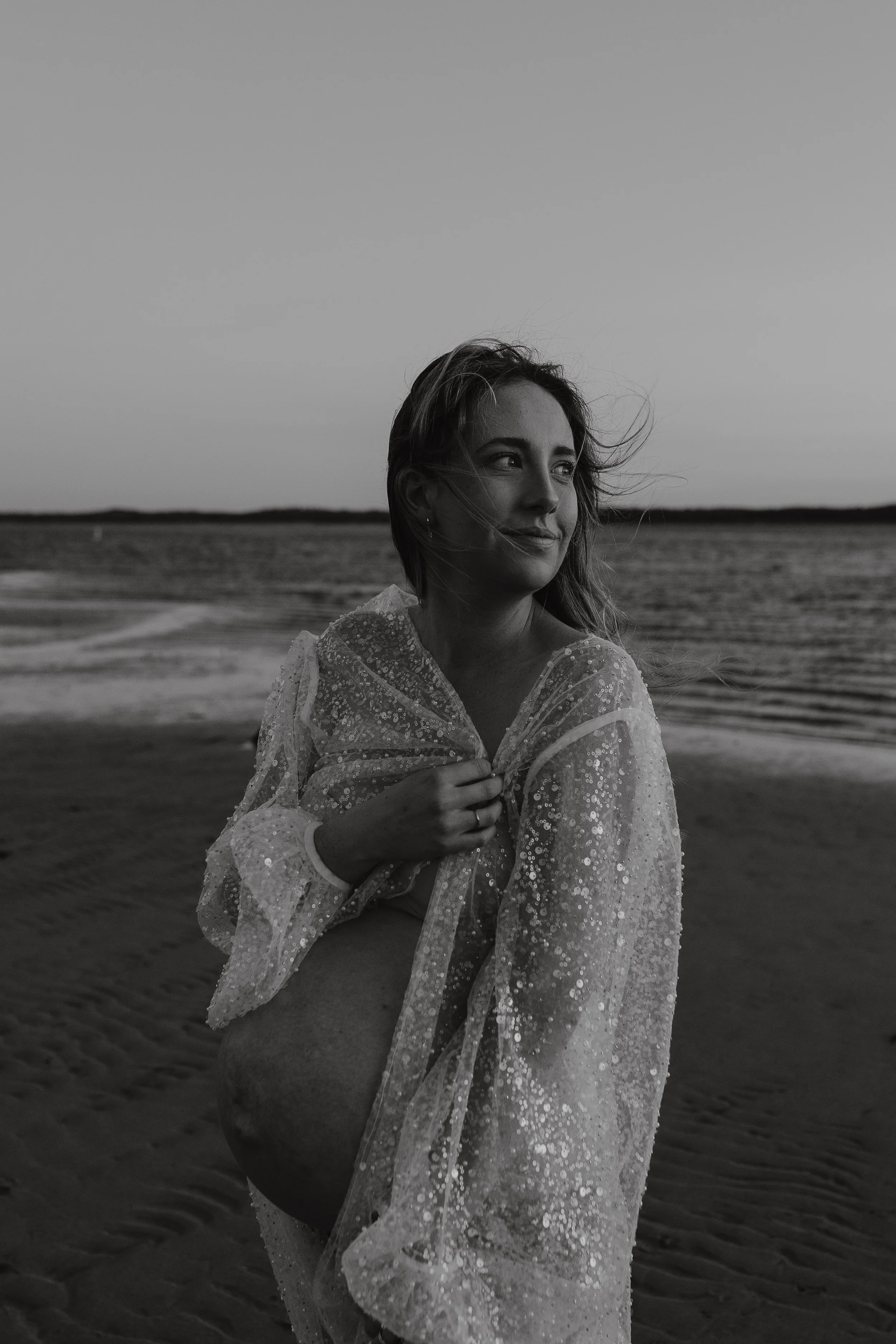 A woman on the beach wearing a sequined, sheer cover-up with wind-blown hair, looking off into the distance with a slight smile, during sunset or dusk.