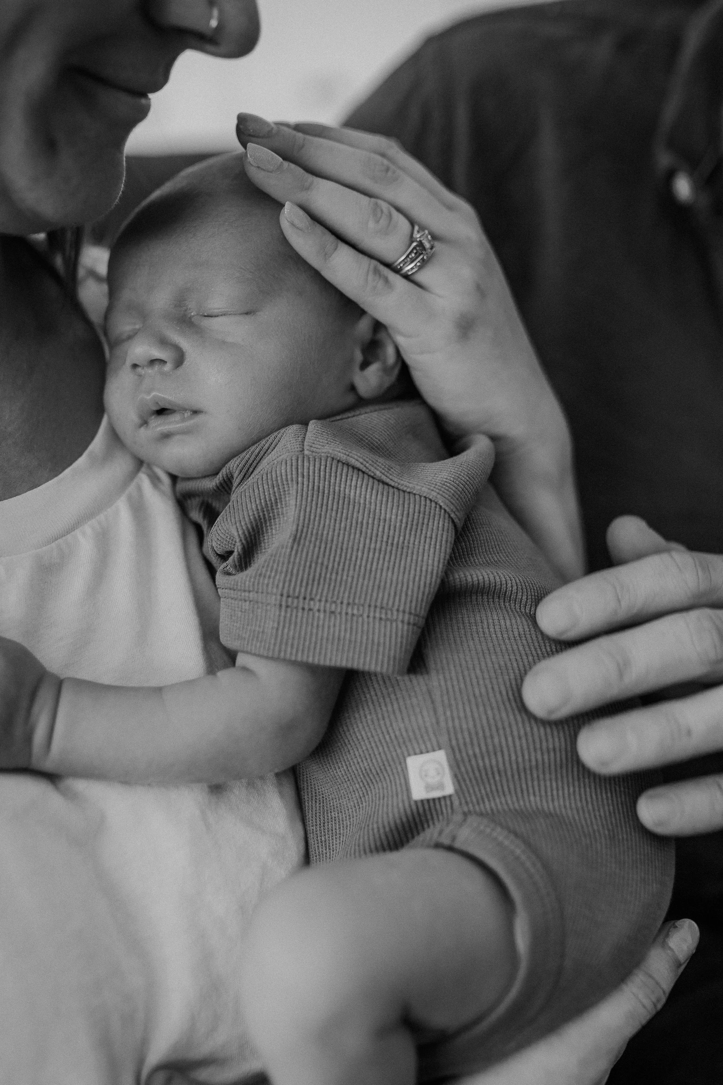 A black and white close-up photo of a person gently holding a sleeping baby with one hand on the baby's forehead and the other supporting the baby's arm, showing a tender moment of care and affection.