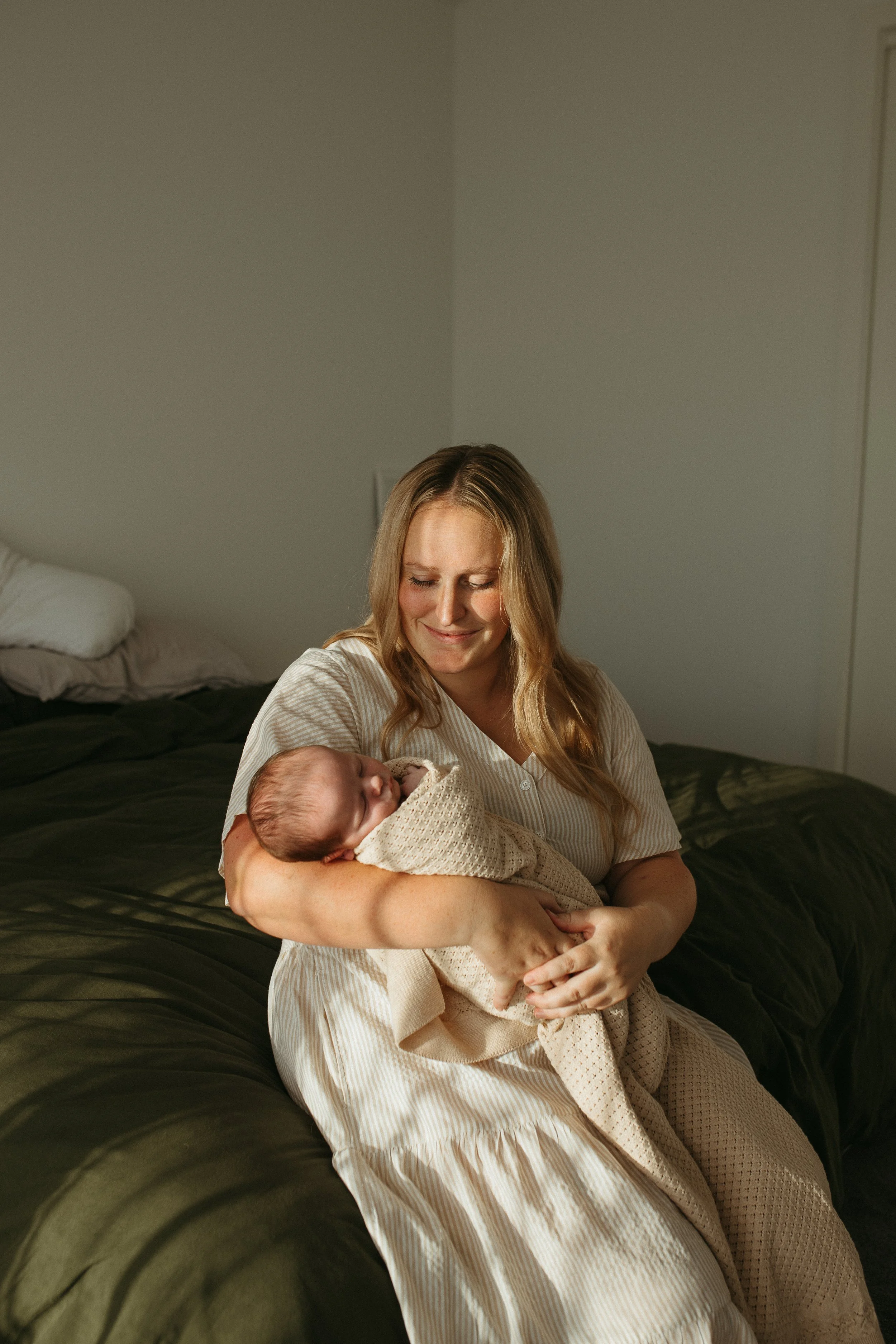 A woman sitting on a bed, holding a sleeping baby wrapped in a beige blanket, smiling gently with closed eyes in a cozy, softly-lit room.