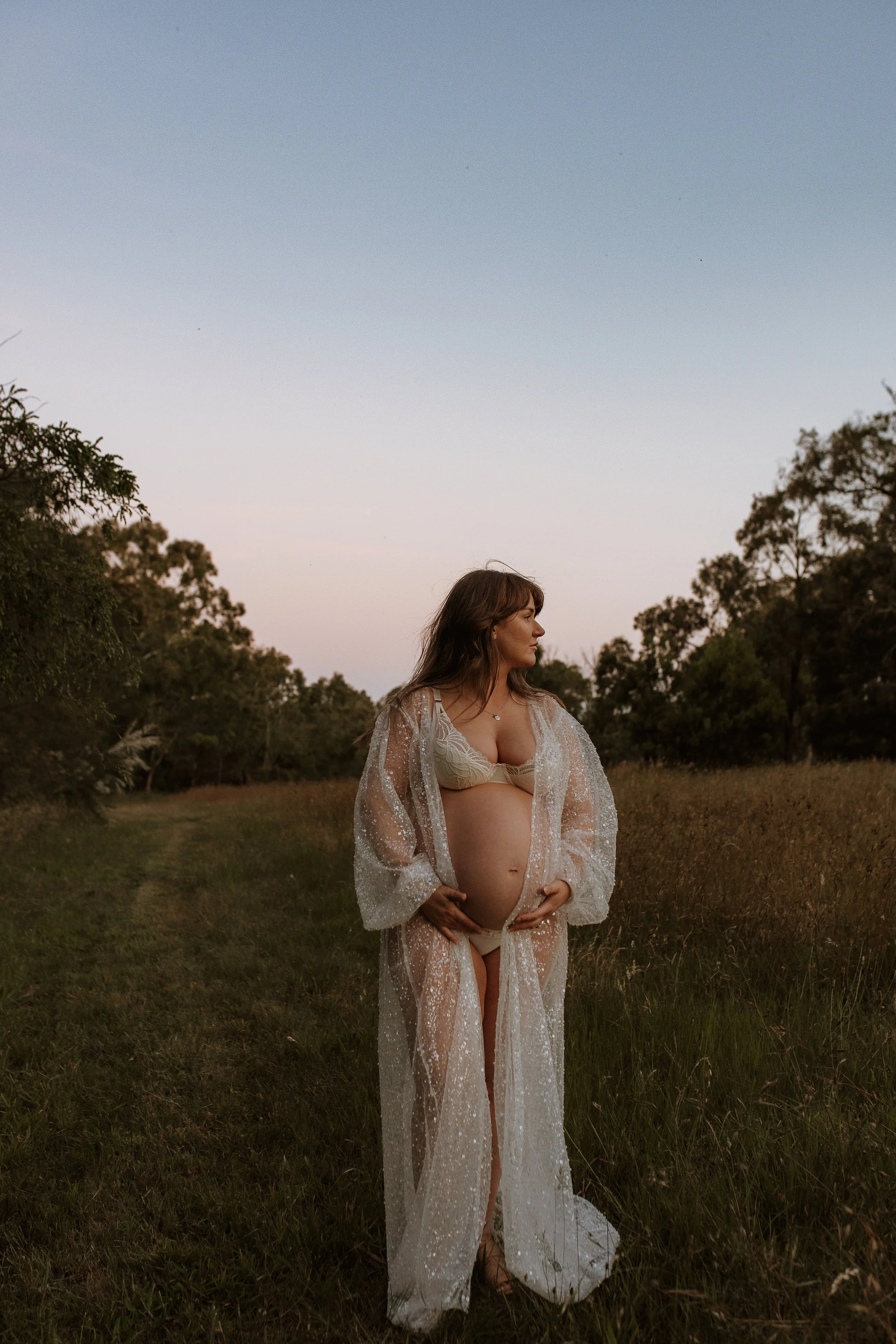 A pregnant woman in a sheer, glittering white robe standing outdoors in a grassy field at dusk, with trees on either side and a clear sky overhead.