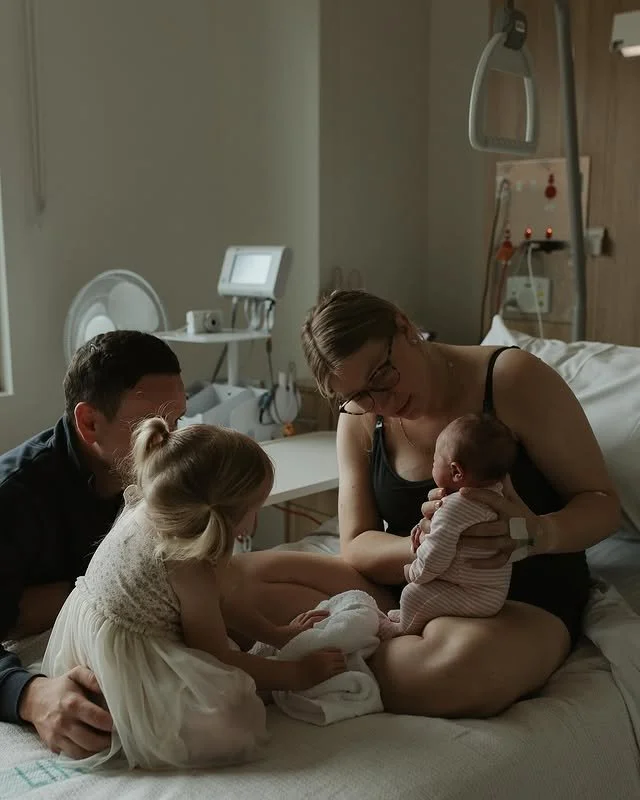 A woman, who appears to be a new mother, is sitting on a hospital bed holding a newborn baby while a man and a young girl sit nearby, all in a hospital room.