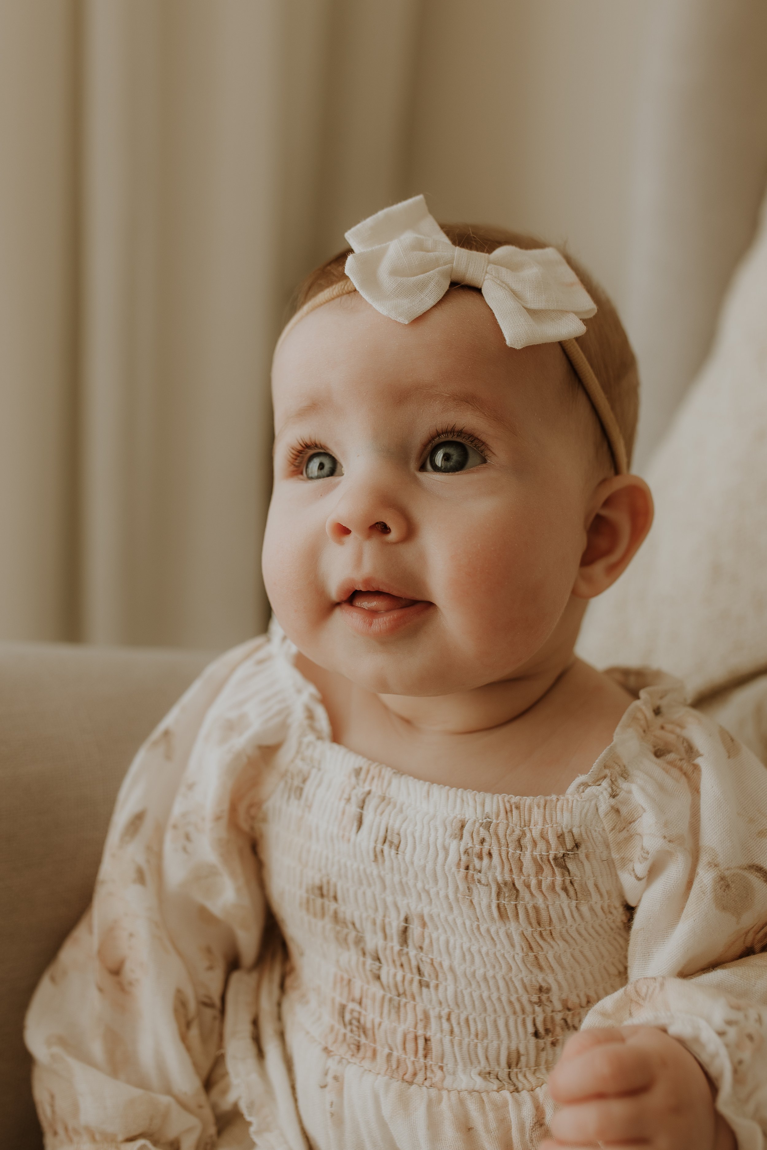 A baby girl with light skin and red hair, wearing a beige top with smocking and a cream-colored bow headband. She is looking up with big blue eyes and her mouth slightly open, sitting on a beige piece of furniture with cream curtains in the background.