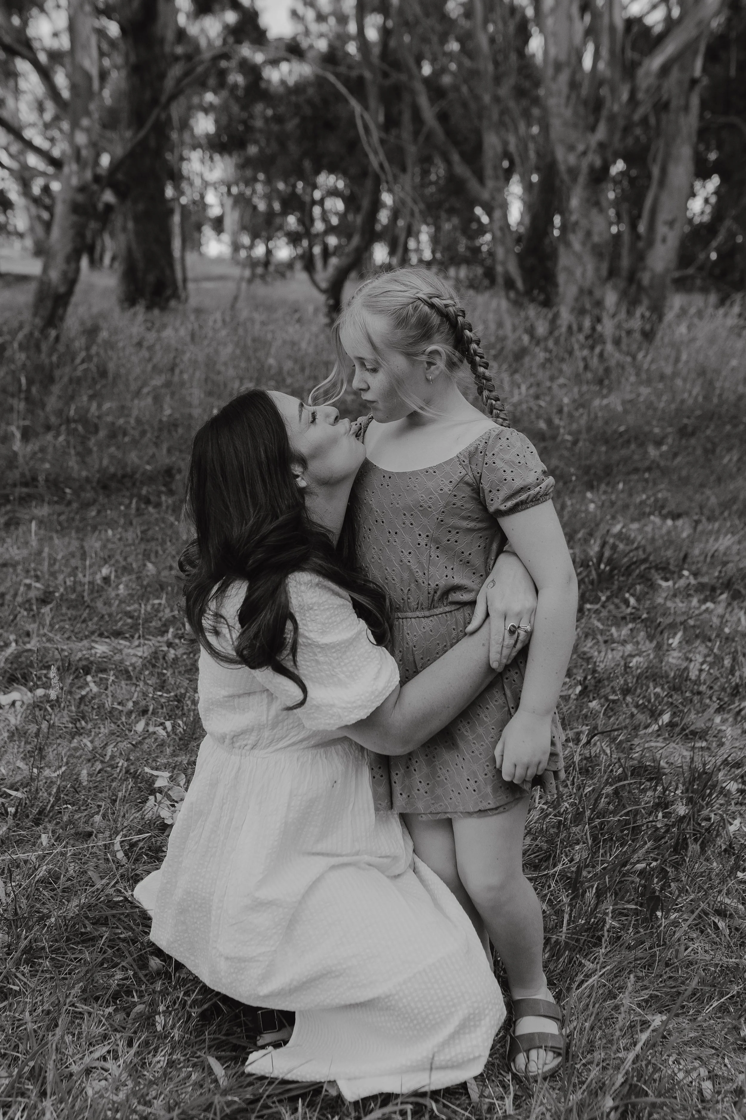 A woman kneeling on the grass and a girl standing in front of her, both looking at each other, in a wooded area.