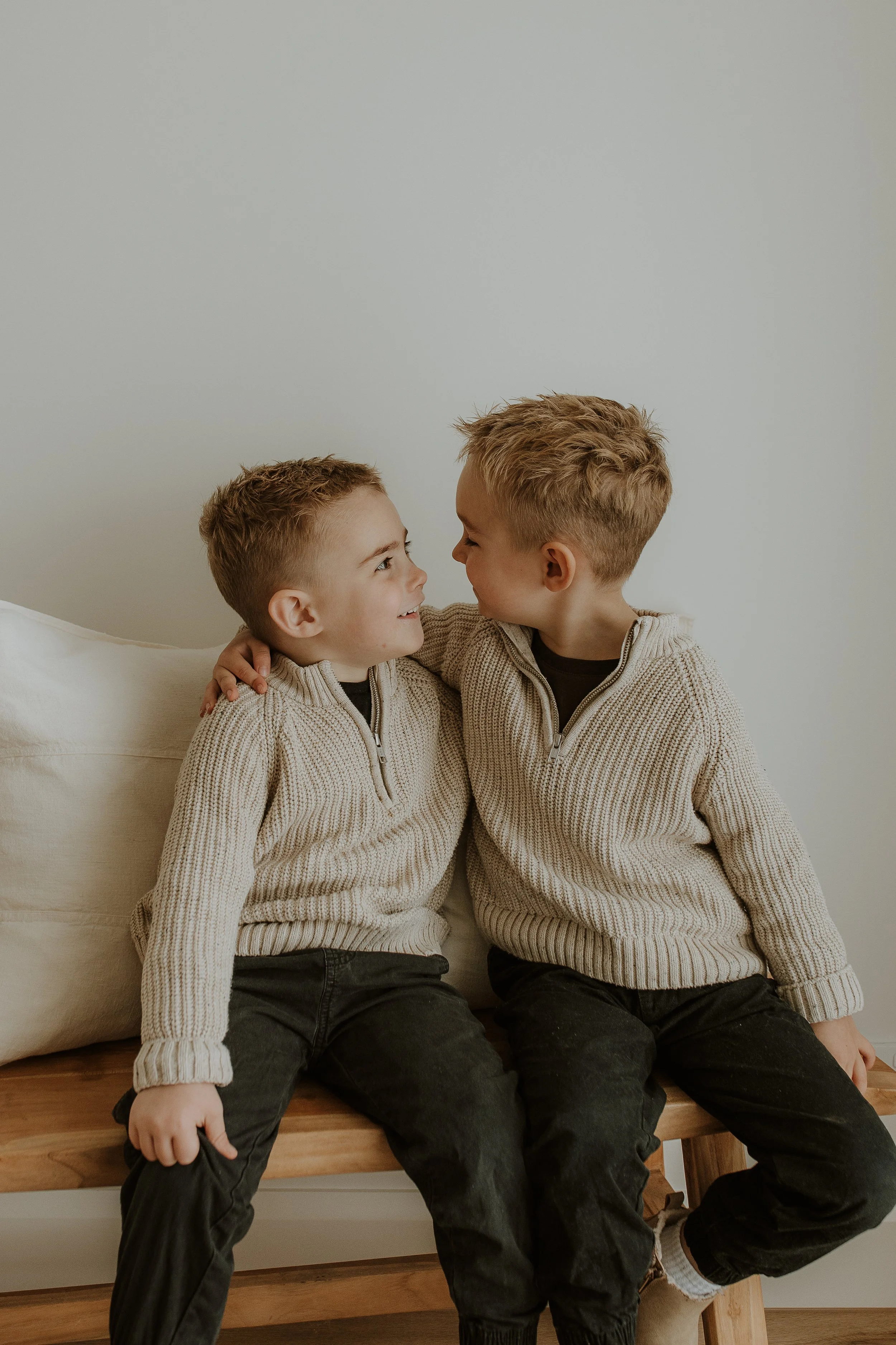Two young boys with short, light brown hair and light skin are sitting on a wooden bench, facing each other, smiling and touching noses. They are wearing matching beige-knit zip-up sweaters and dark pants, with one boy's hand on the other's shoulder.