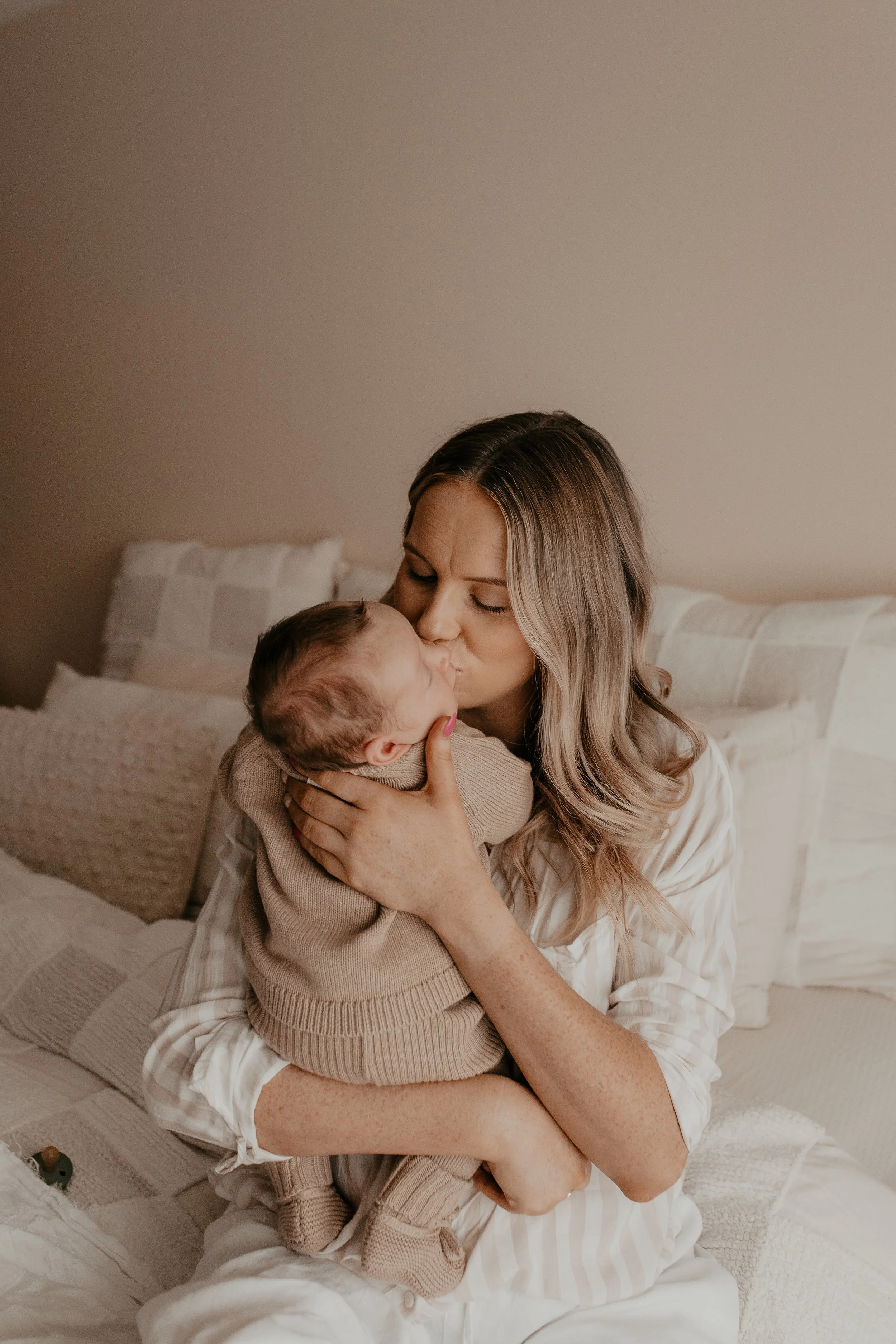 A woman with long blonde hair and freckles is sitting on a bed, holding and kissing a sleeping baby wrapped in a beige sweater. The background shows a beige wall and a white bed with beige pillows.
