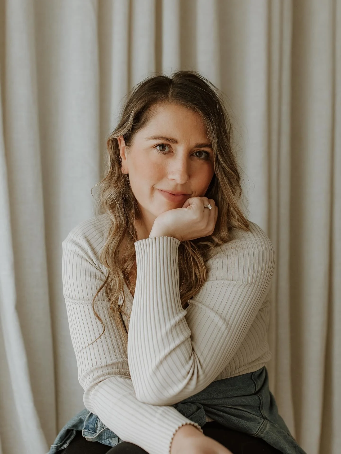 A woman with long, wavy hair and light skin, sitting in front of a beige curtain, wearing a cream ribbed sweater, resting her chin on her hand and smiling softly.