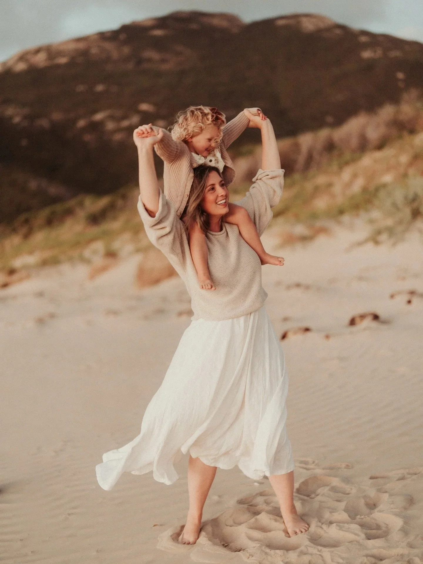 A woman with a long white skirt and a beige sweater carrying a girl with curly blonde hair on her shoulders in a sandy outdoor setting with hills in the background.